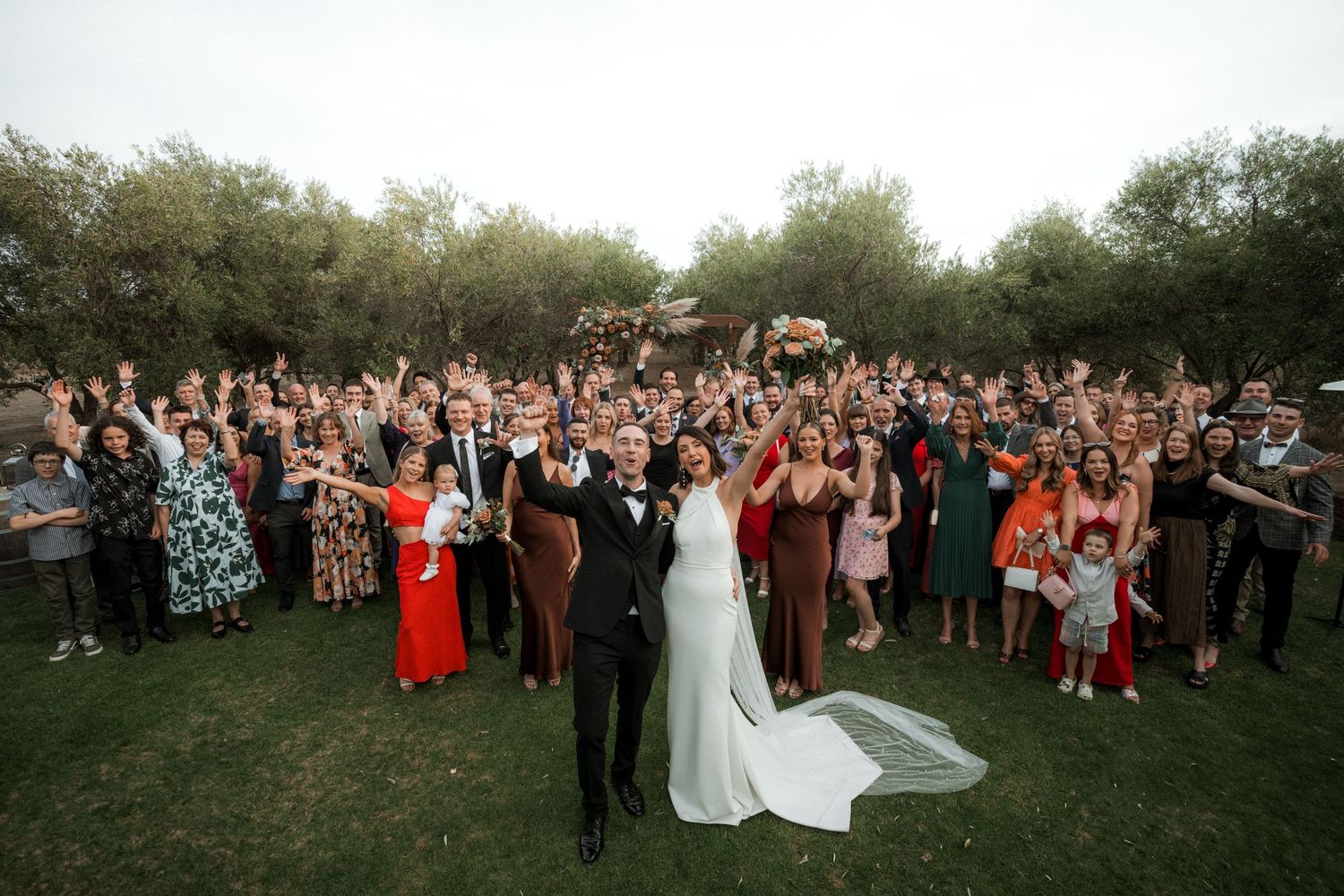 Large outdoor wedding reception crowd celebrates with couple on grassy lawn during sunset.