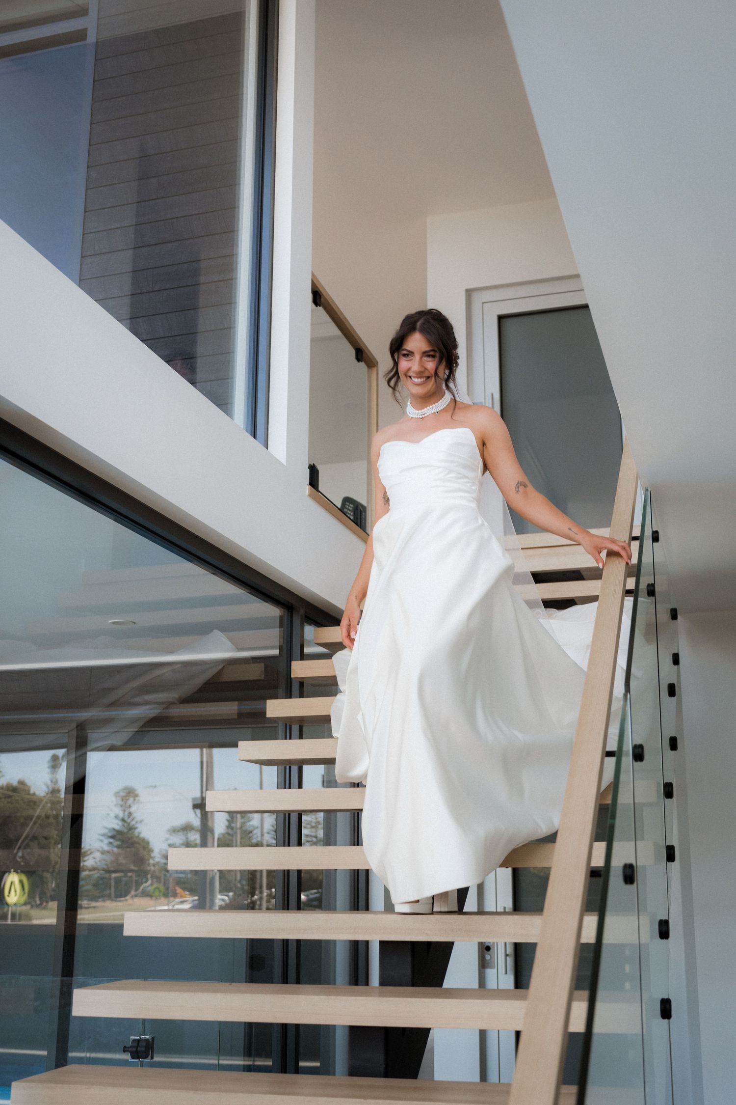 A bride in a flowing white wedding dress descends a modern floating staircase in an elegant indoor setting.