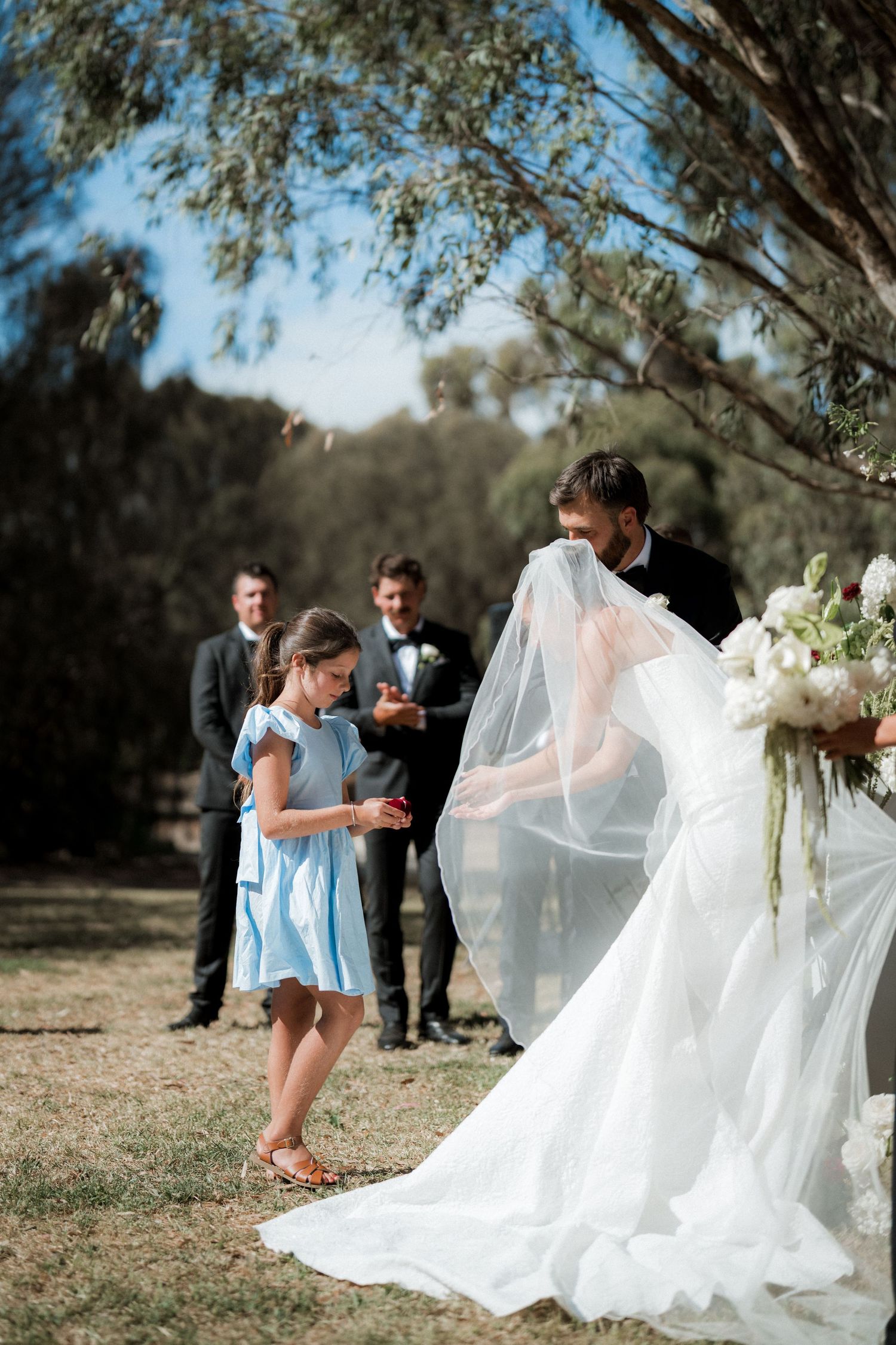 Groom lifting bride's veil as a young girl in blue dress watches during an outdoor wedding ceremony.
