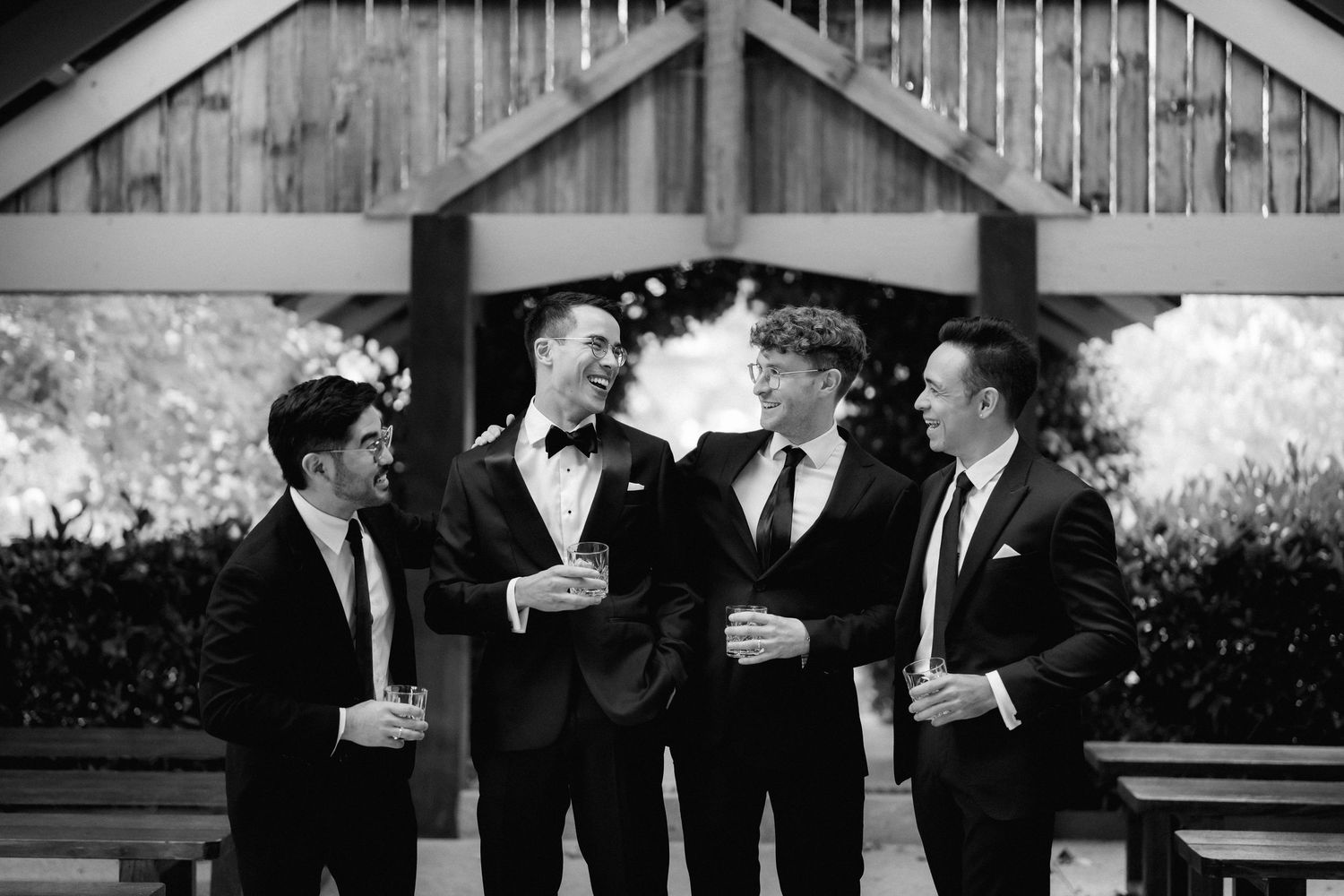 Black and white photo of groom and groomsmen in tuxedos laughing together outdoors at a wedding venue.
