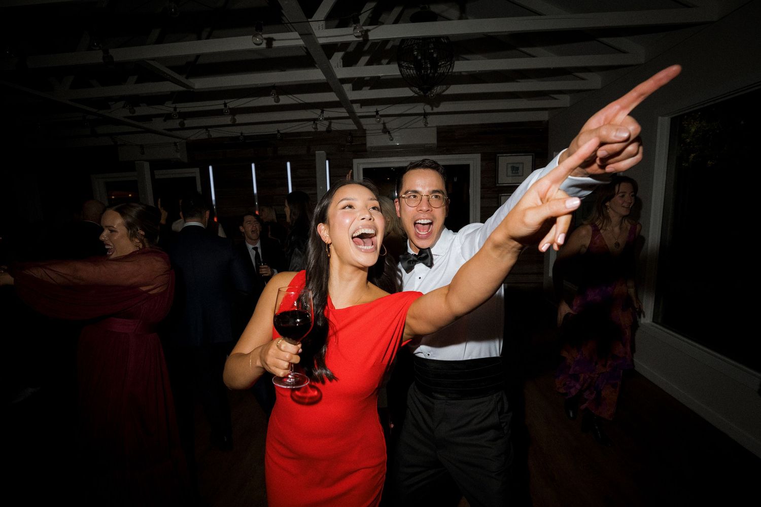 Excited wedding guests in red and white dancing and pointing at indoor reception celebration.