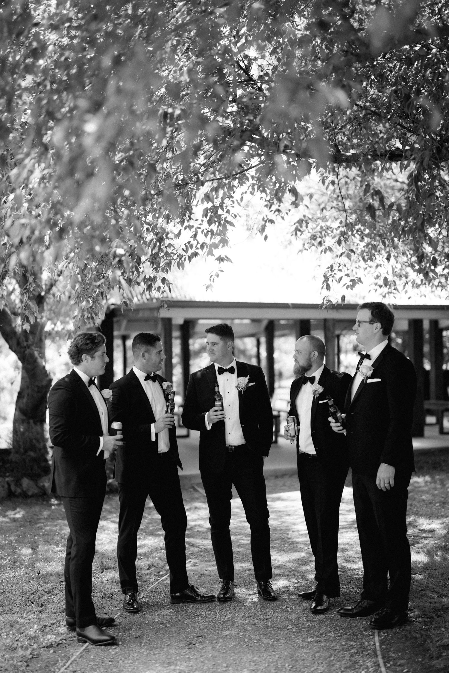Group of groomsmen in black tuxedos having drinks and conversation under a tree at an outdoor wedding.
