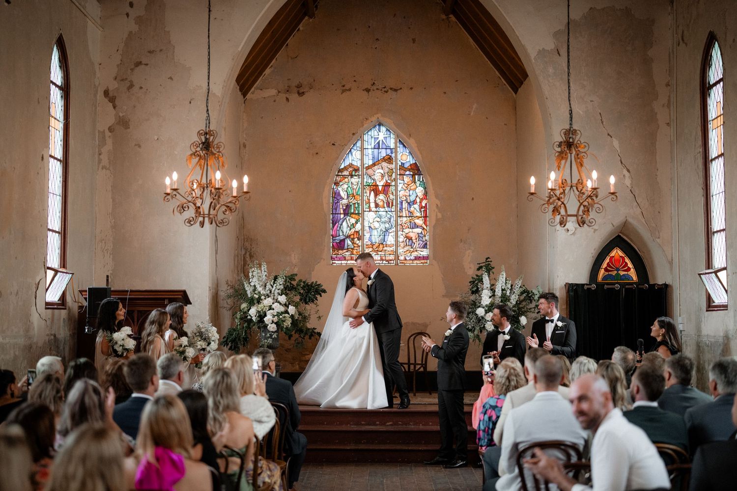 A couple shares an intimate moment during their wedding ceremony inside a historic chapel with stained glass windows.