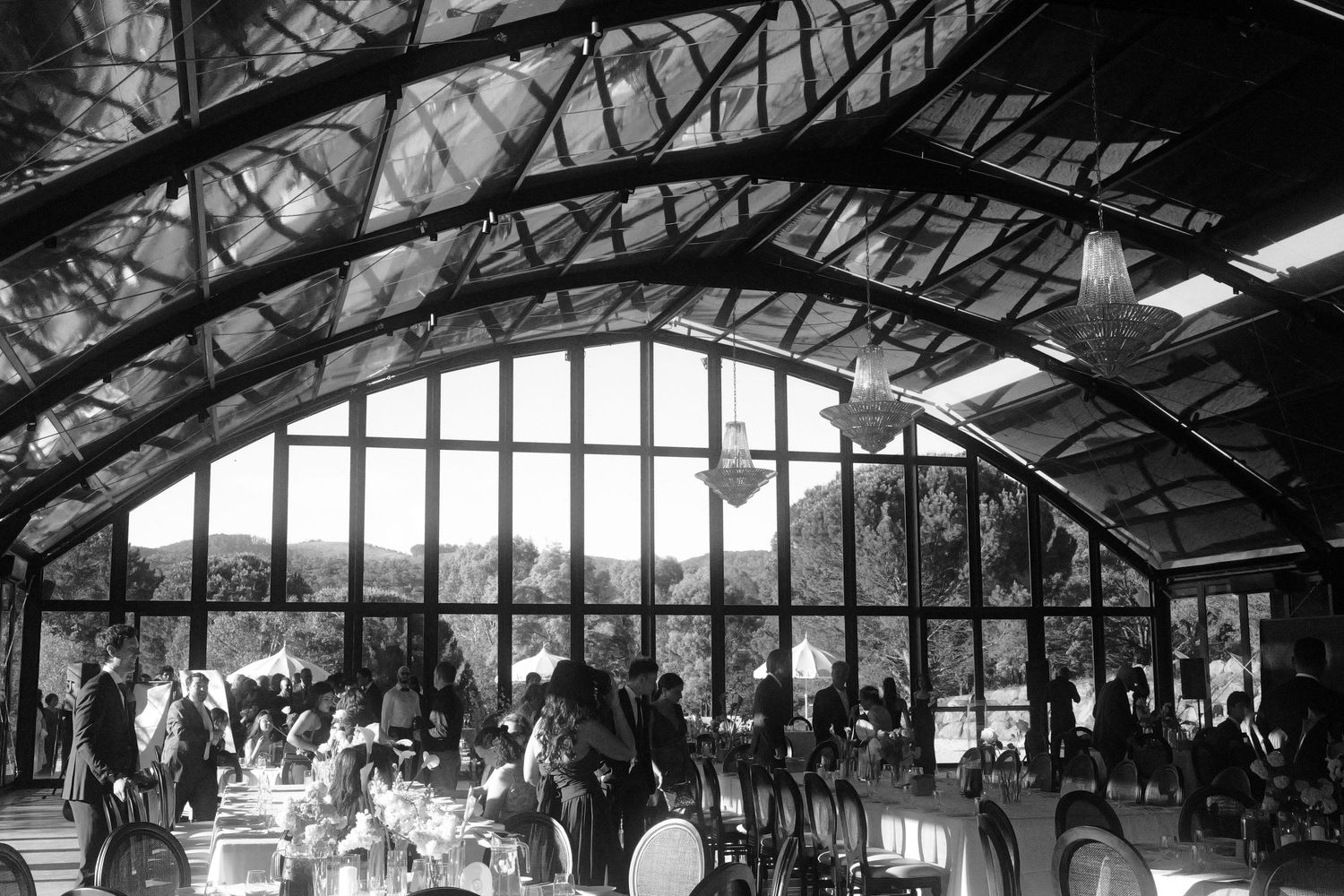 Black and white view of wedding guests seated at tables inside a bright glass-walled pavilion with an arched wooden ceiling and chandeliers.