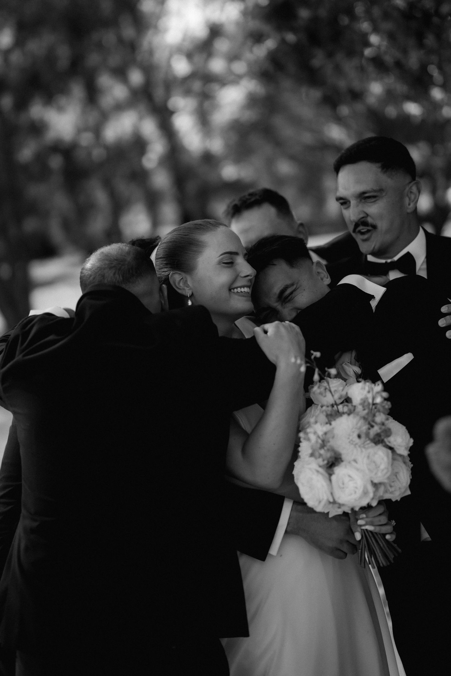 A black and white wedding party photo showing emotional embraces and bouquets of white flowers.