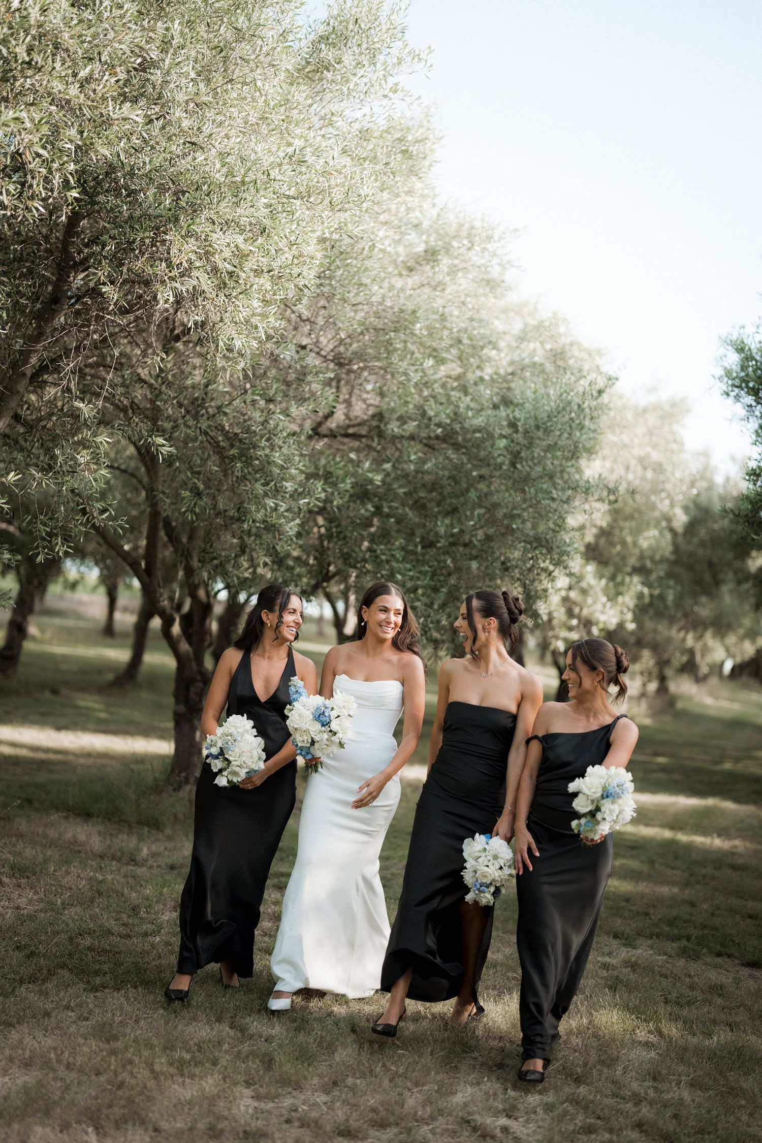 Wedding party walking through an orchard with bridesmaids in black dresses and white bouquets.