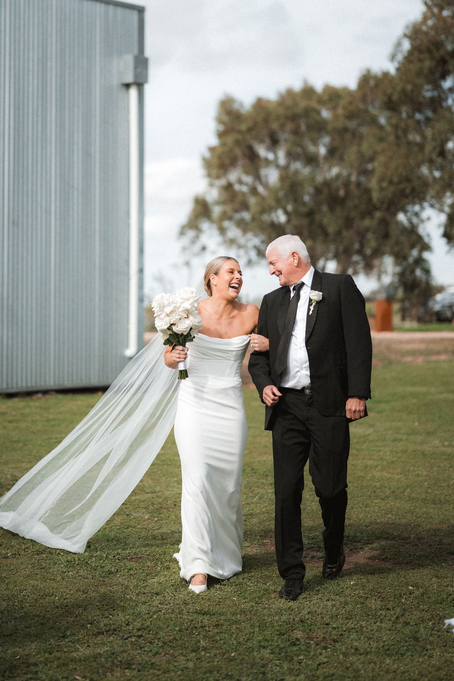 Wedding couple walks and poses playfully near industrial building with flowing veil in the wind.