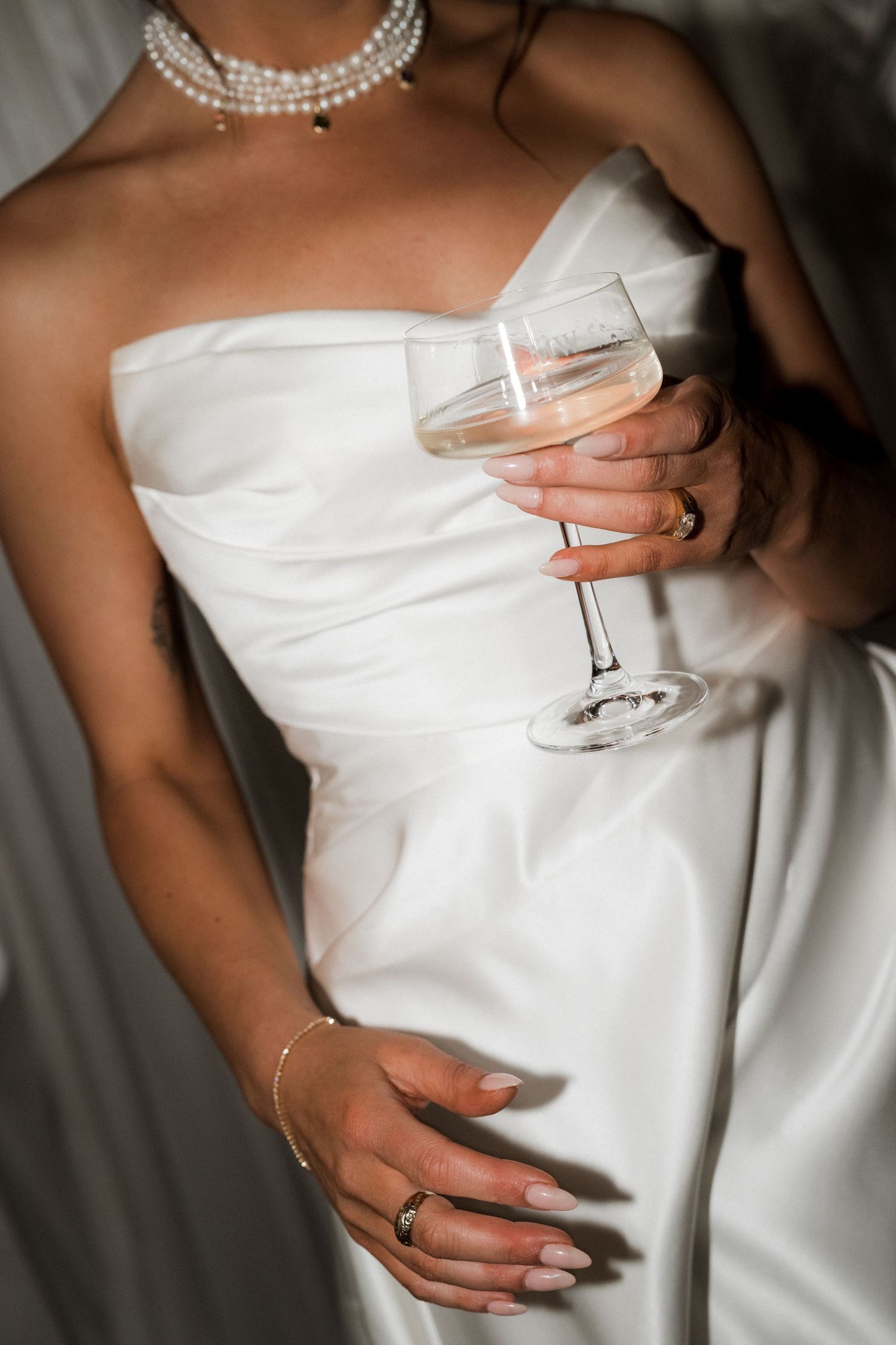 Elegant white wedding dress detail with pearl necklace and champagne glass held by the bride.