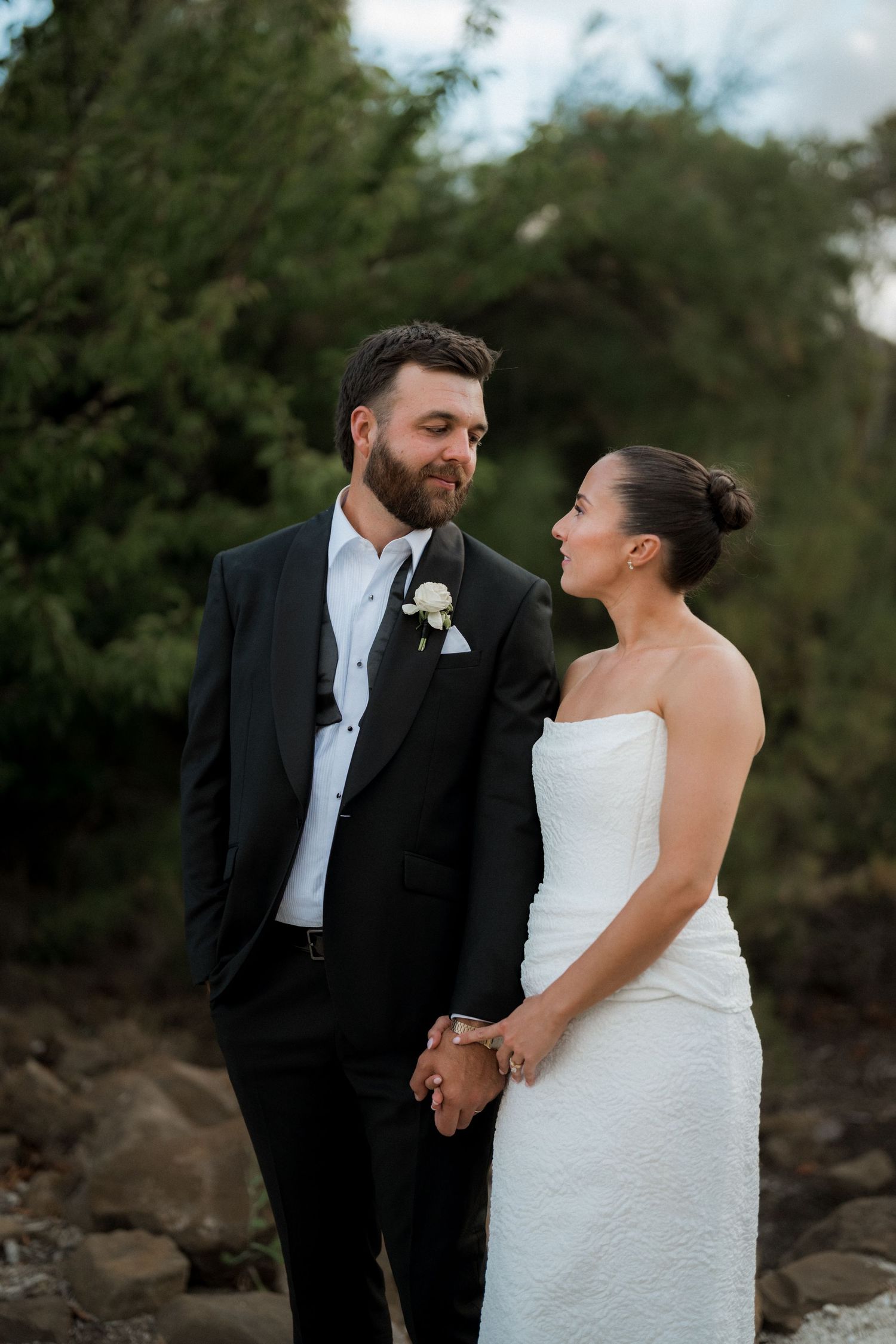 Bride and groom holding hands and gazing at each other outdoors, she in strapless white gown, he in black suit.