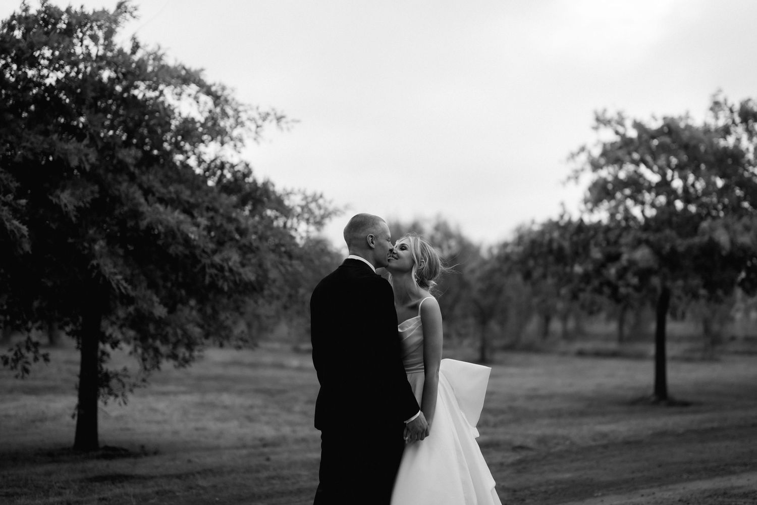 Black and white photo of newlyweds embracing in an orchard setting.