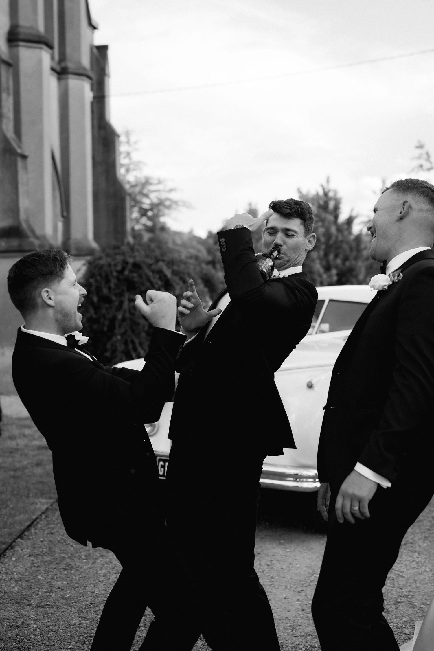 A vintage black and white photo showing groomsmen in suits laughing and joking together outside.