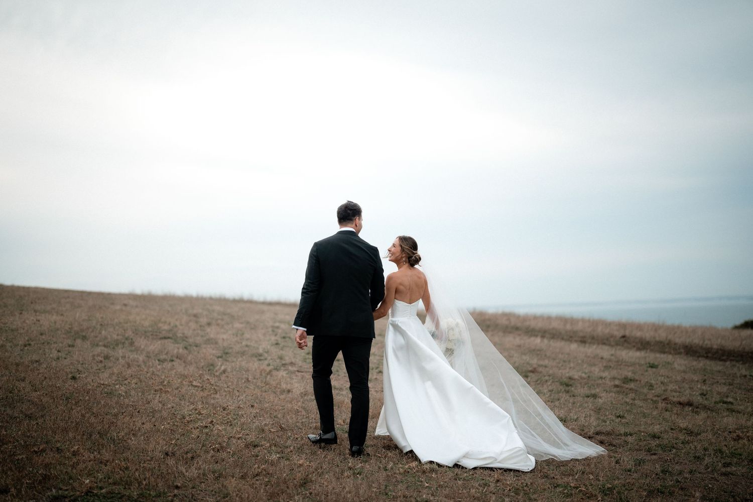 A newlywed couple walking together on a hilltop against an overcast sky, with the bride wearing a flowing white gown.