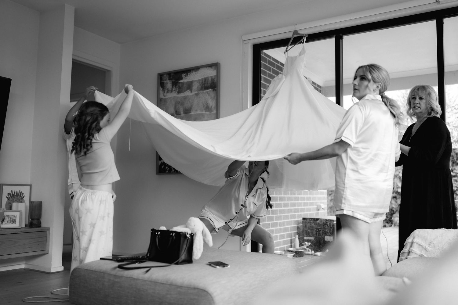 Black and white photo of people preparing and holding up a wedding dress in a bright indoor room.