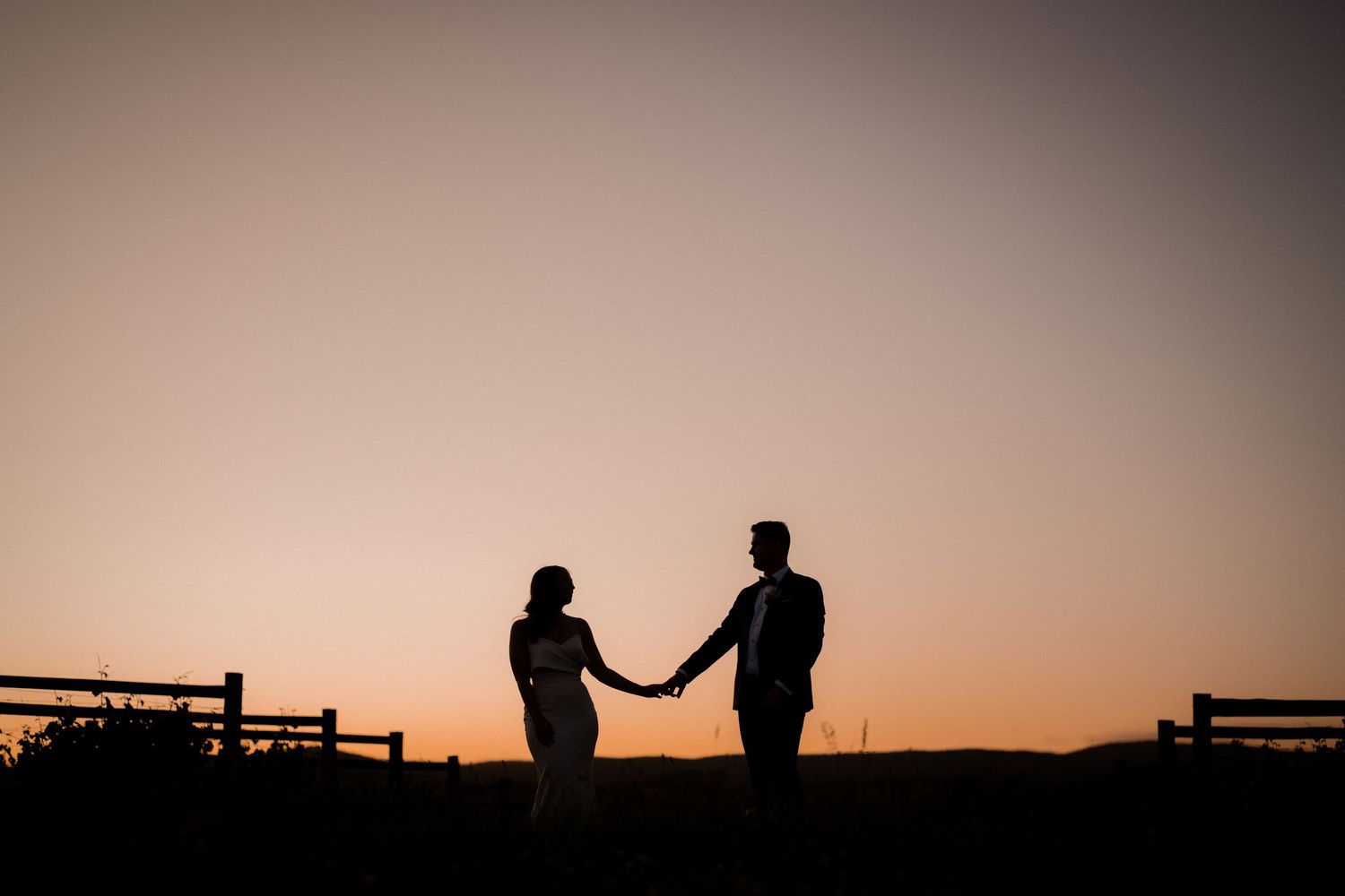 Silhouetted couple holding hands at sunset against a rustic fence backdrop in a romantic countryside setting.