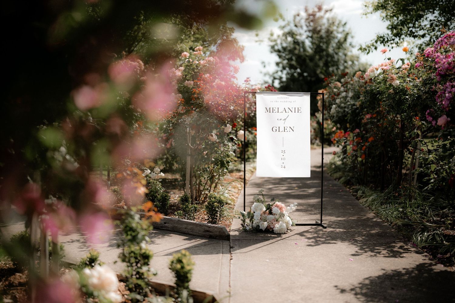 A romantic garden path with pink blooming flowers leads to an elegant white welcome sign in dappled sunlight.
