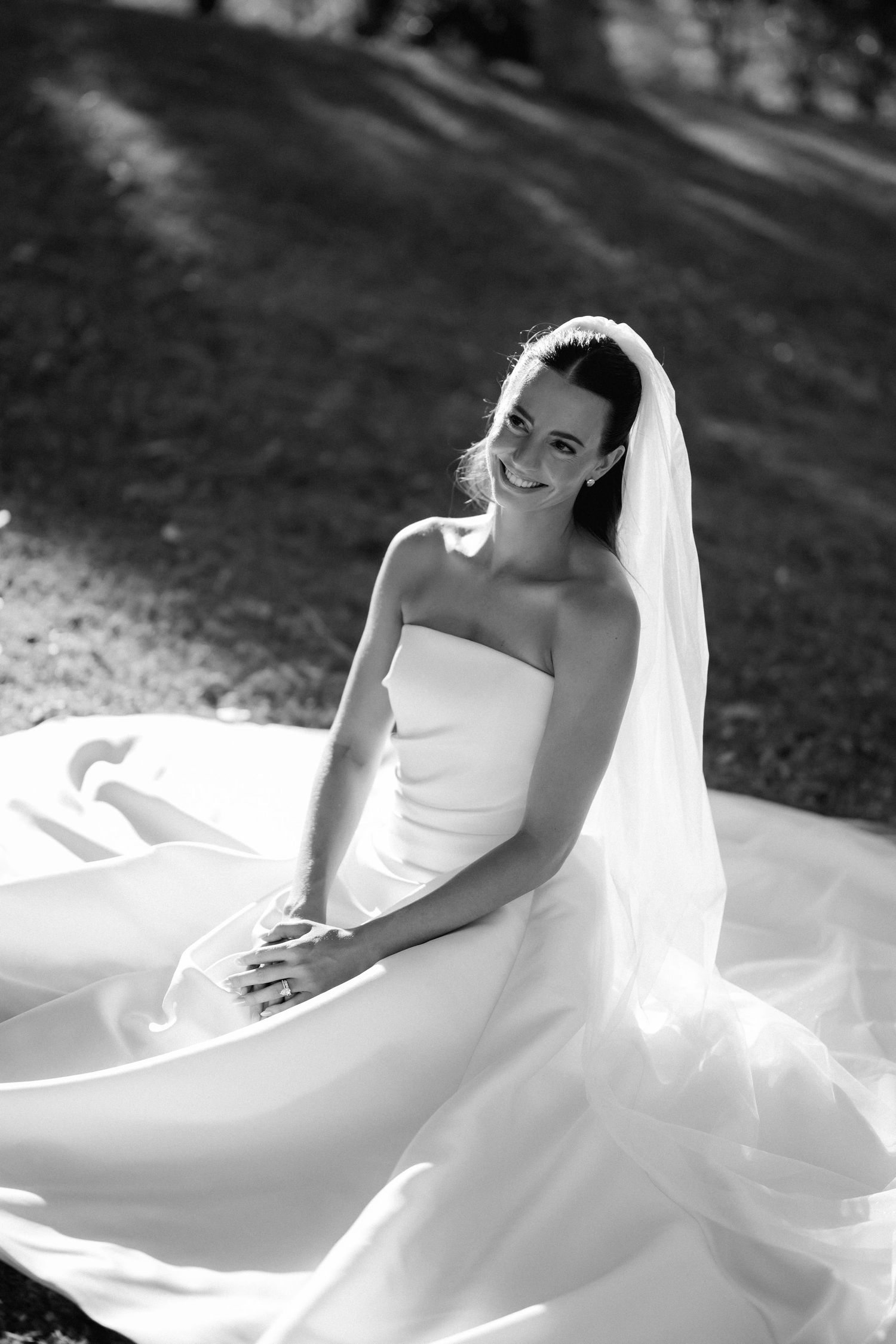 Black and white portrait of bride in flowing white wedding dress sitting gracefully outdoors.