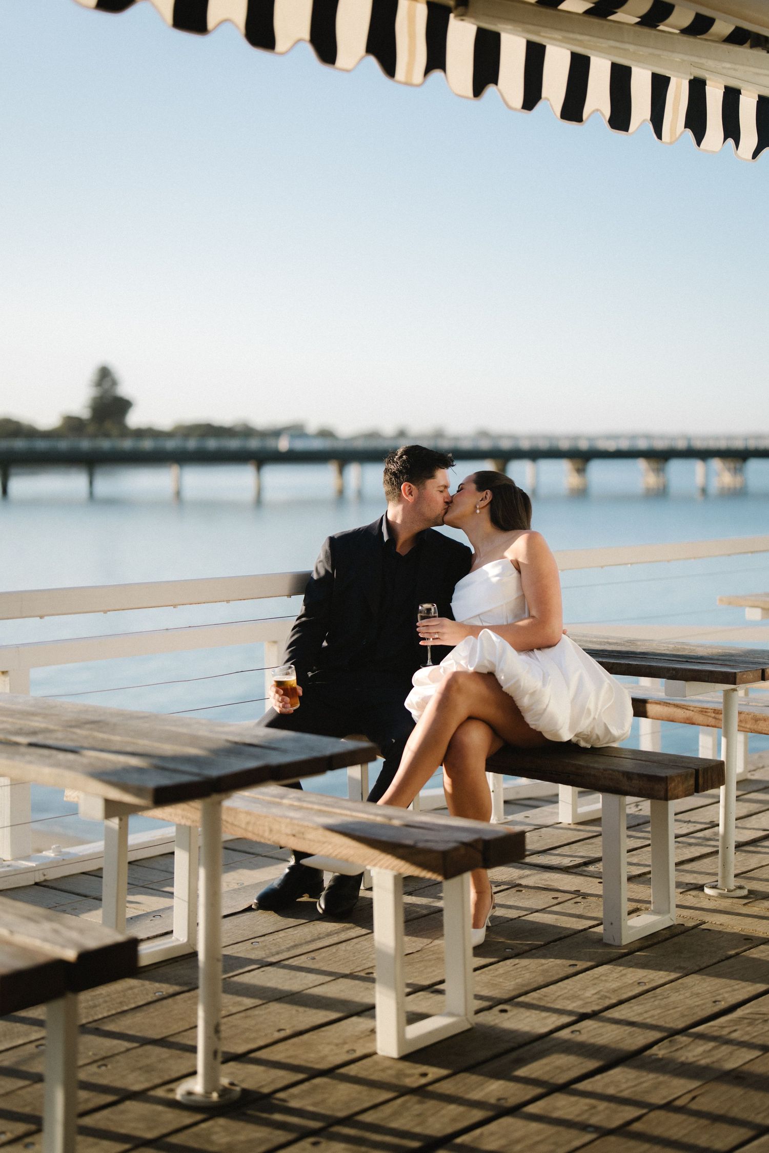 Two people sit together on a wooden bench at a pier during sunset casting long shadows on the boardwalk.