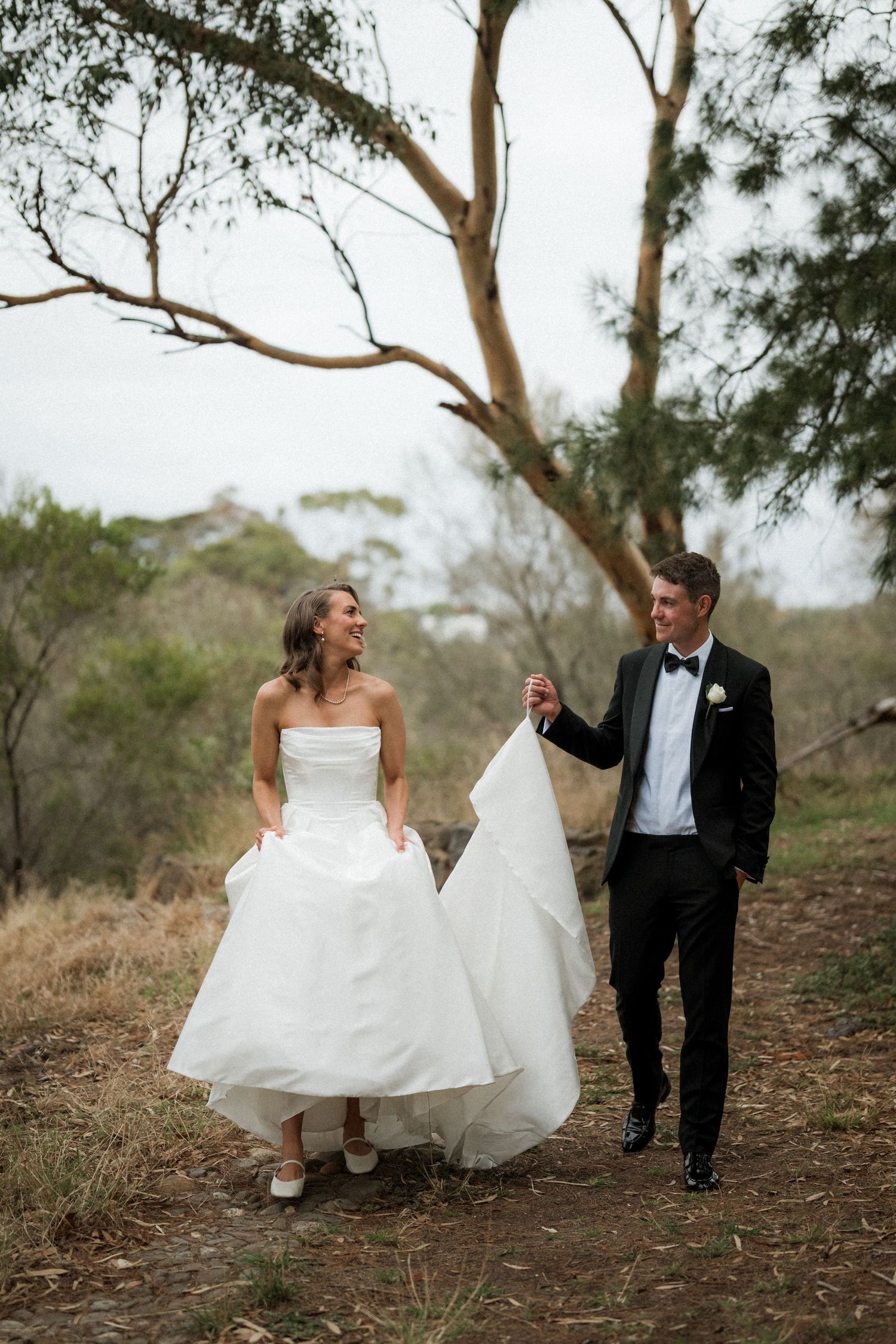 Couple walking playfully through trees in wedding attire.