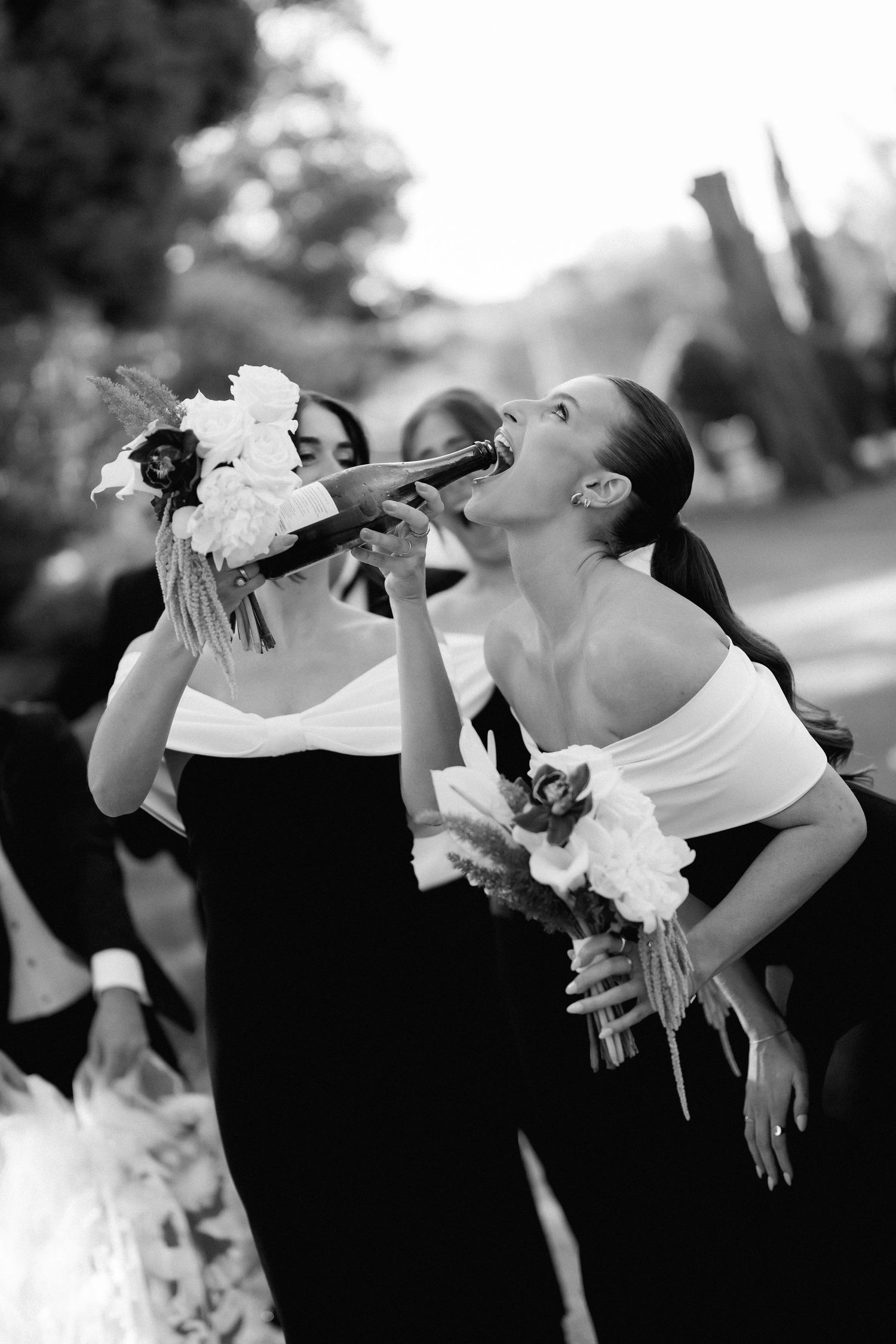 Bridesmaids in black and white attire drink champagne while holding bouquets during wedding celebrations.