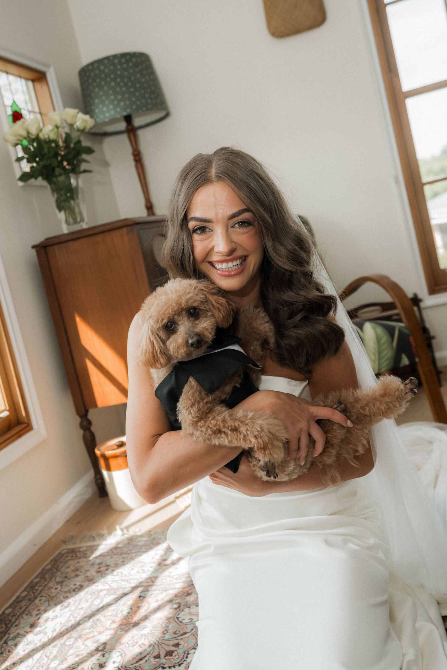 A person in a white dress cuddles with a small brown dog in a sunlit room with green decor.