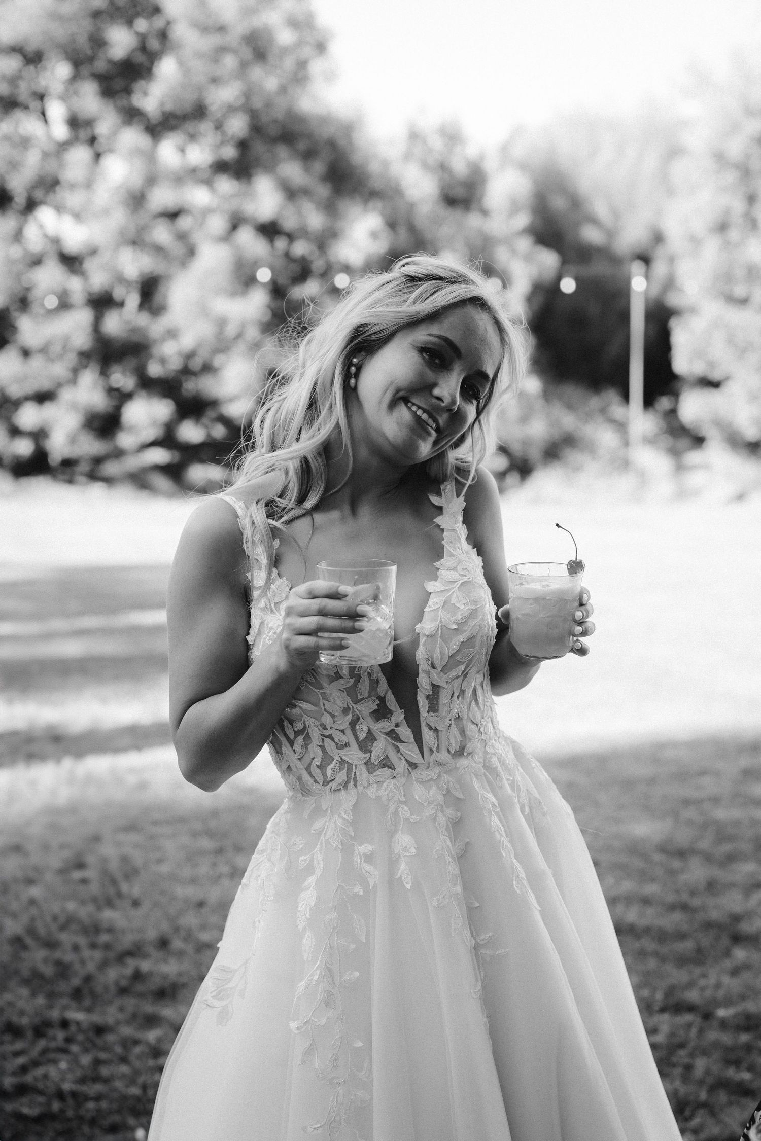 A joyful bride in a lace wedding dress holds two drinks while enjoying a sunny outdoor celebration.