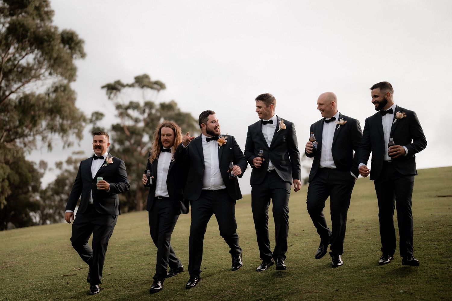 Groomsmen in matching black tuxedos walk together on a grassy hillside during a wedding photoshoot.