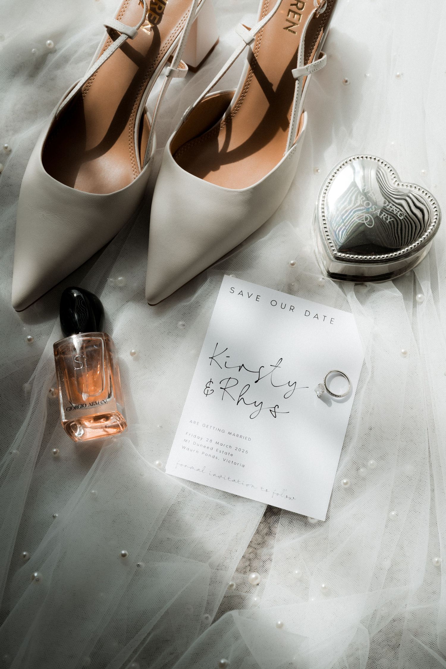 Wedding details flatlay with beige heels, perfume bottle, heart-shaped trinket box and save the date card on marble surface.