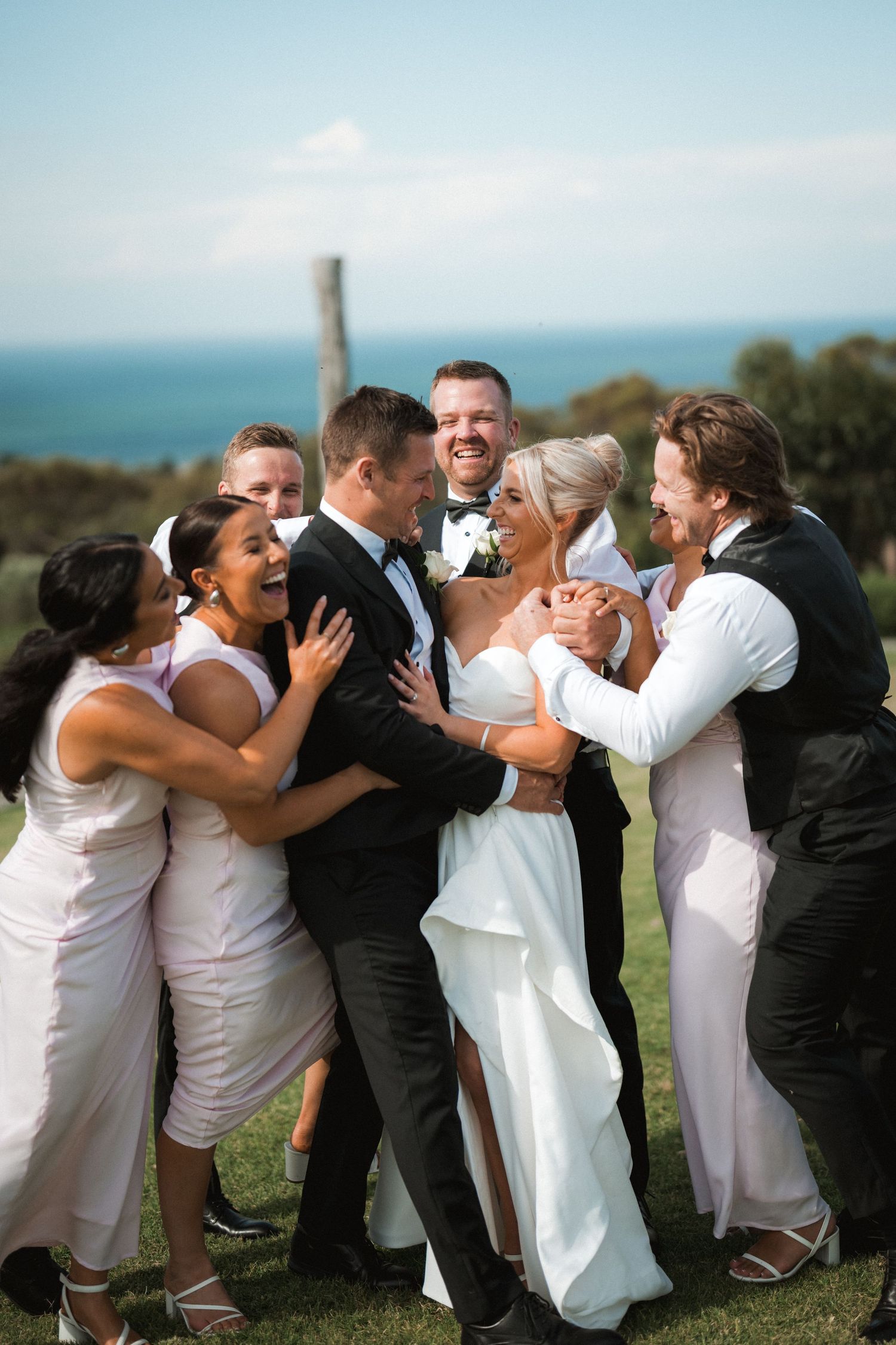 Wedding party members share a joyful group hug outdoors with ocean views.