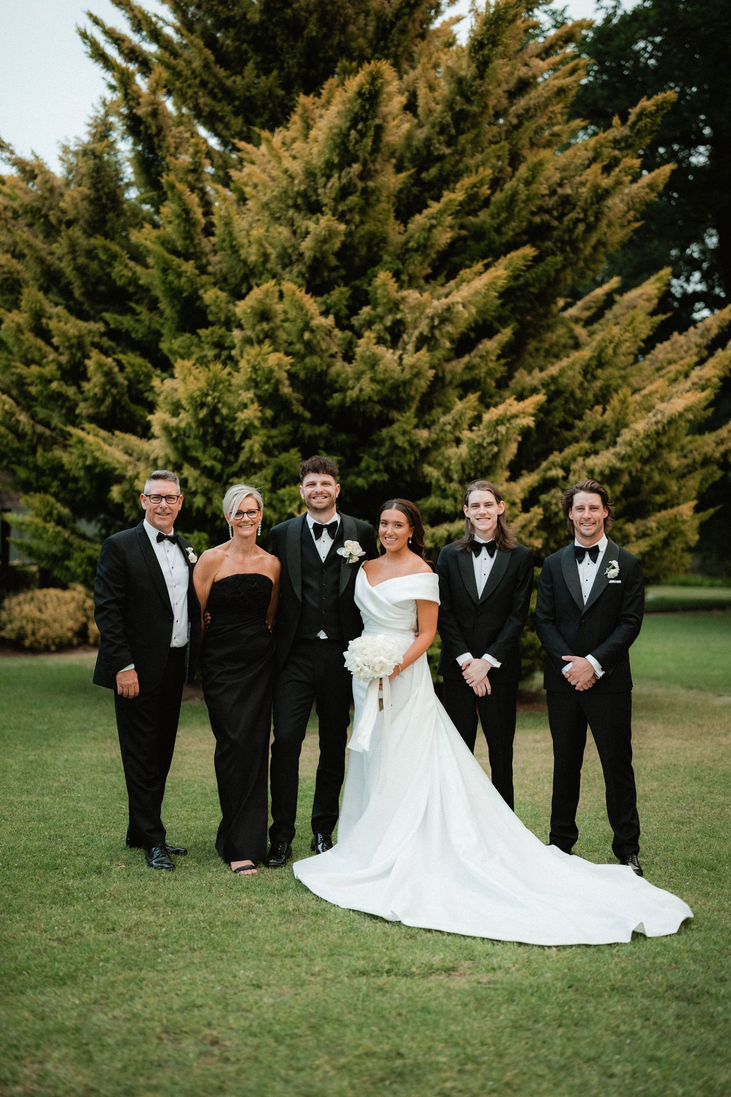 Wedding party poses for formal photo in front of evergreen trees on a manicured lawn.