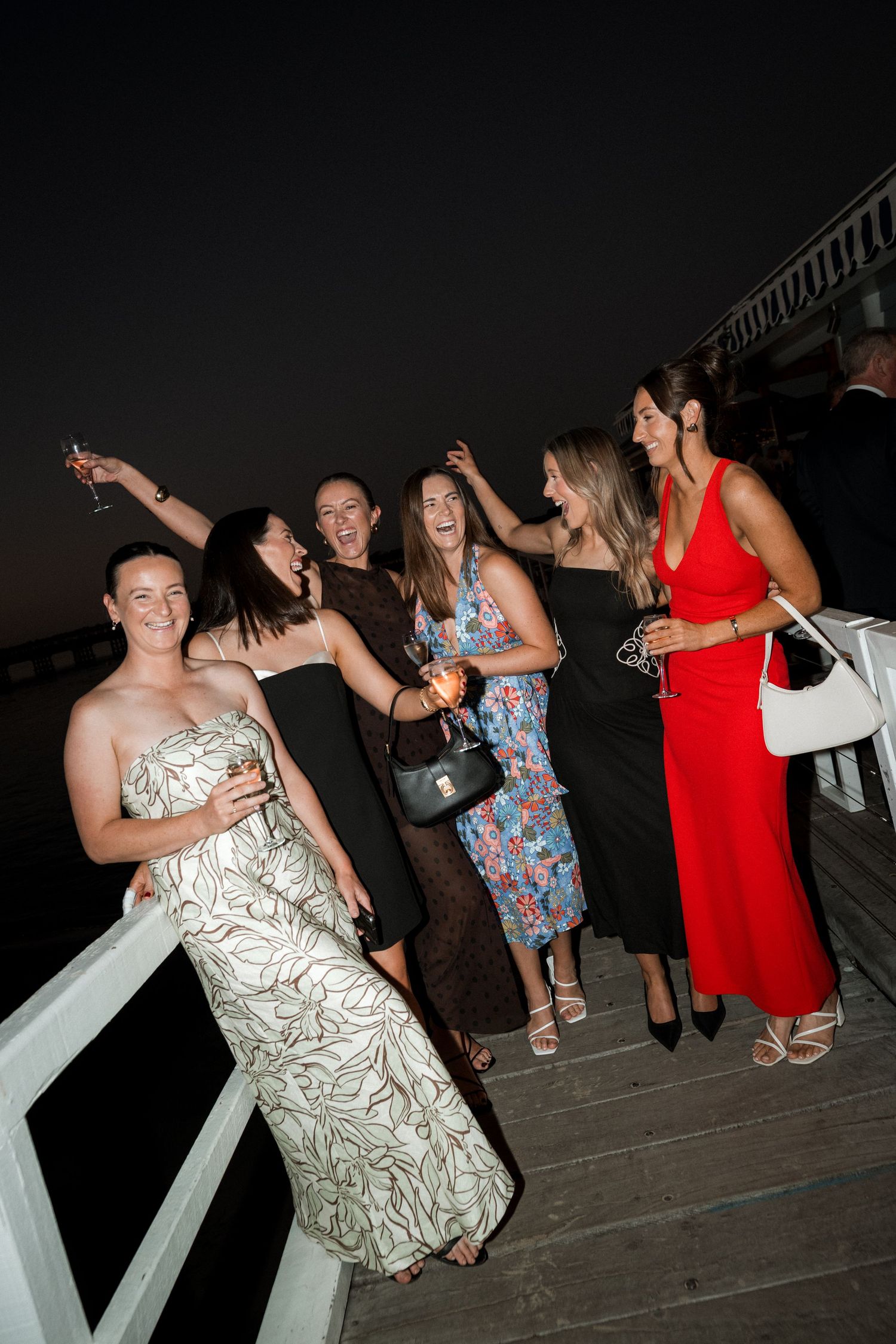 A group of elegantly dressed guests celebrate on a dark wooden deck at night.