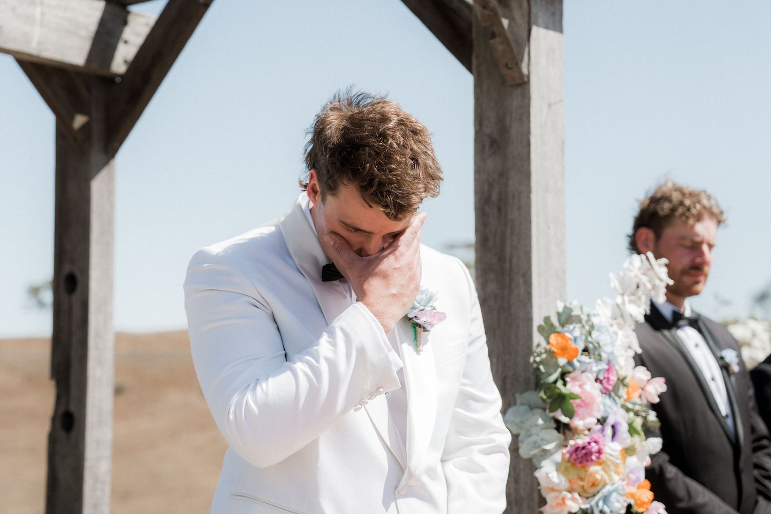 Emotional groom in white suit wiping tears at outdoor wedding ceremony altar.