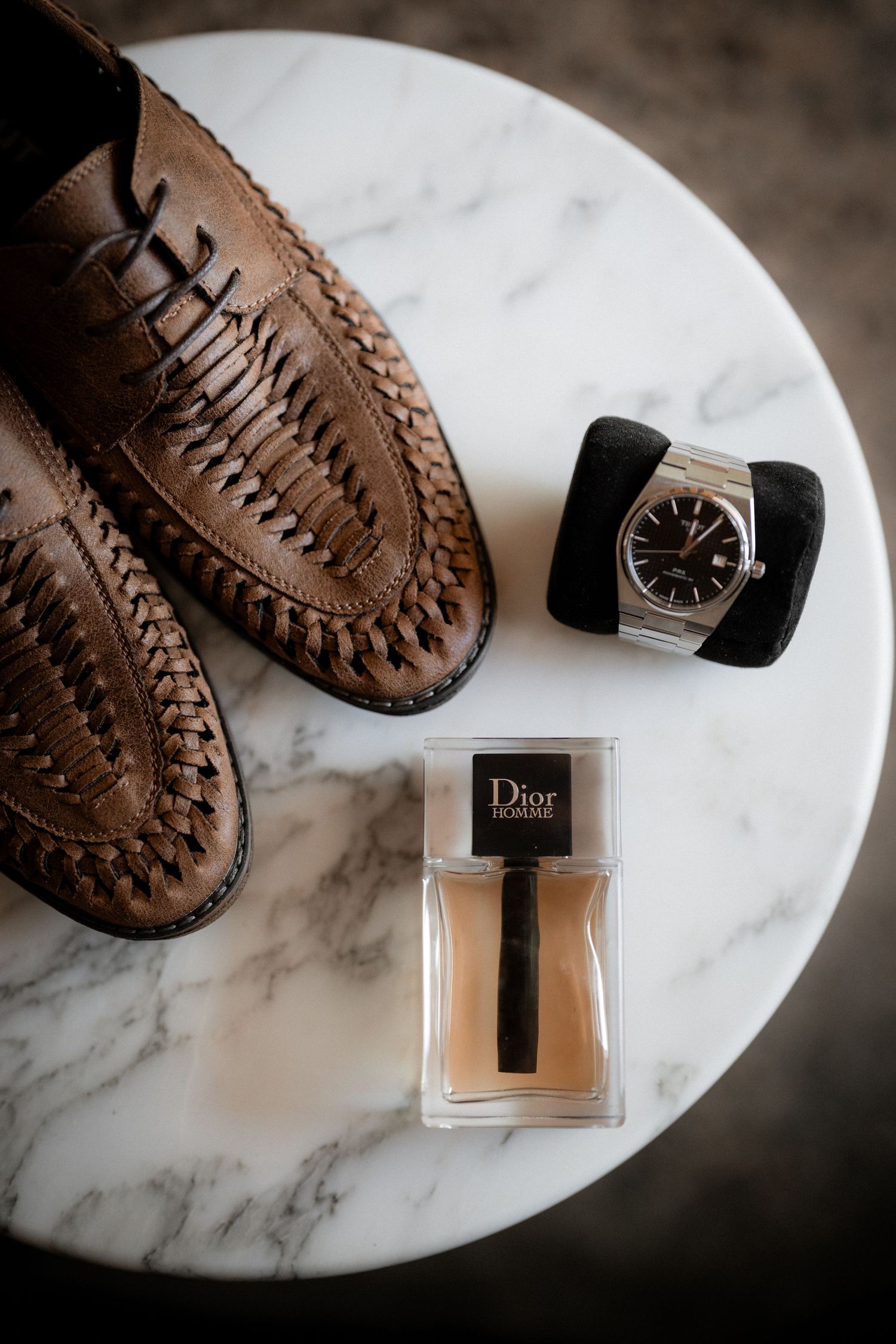 Brown leather shoes and Dior perfume bottle arranged on white marble surface for luxury product photography.