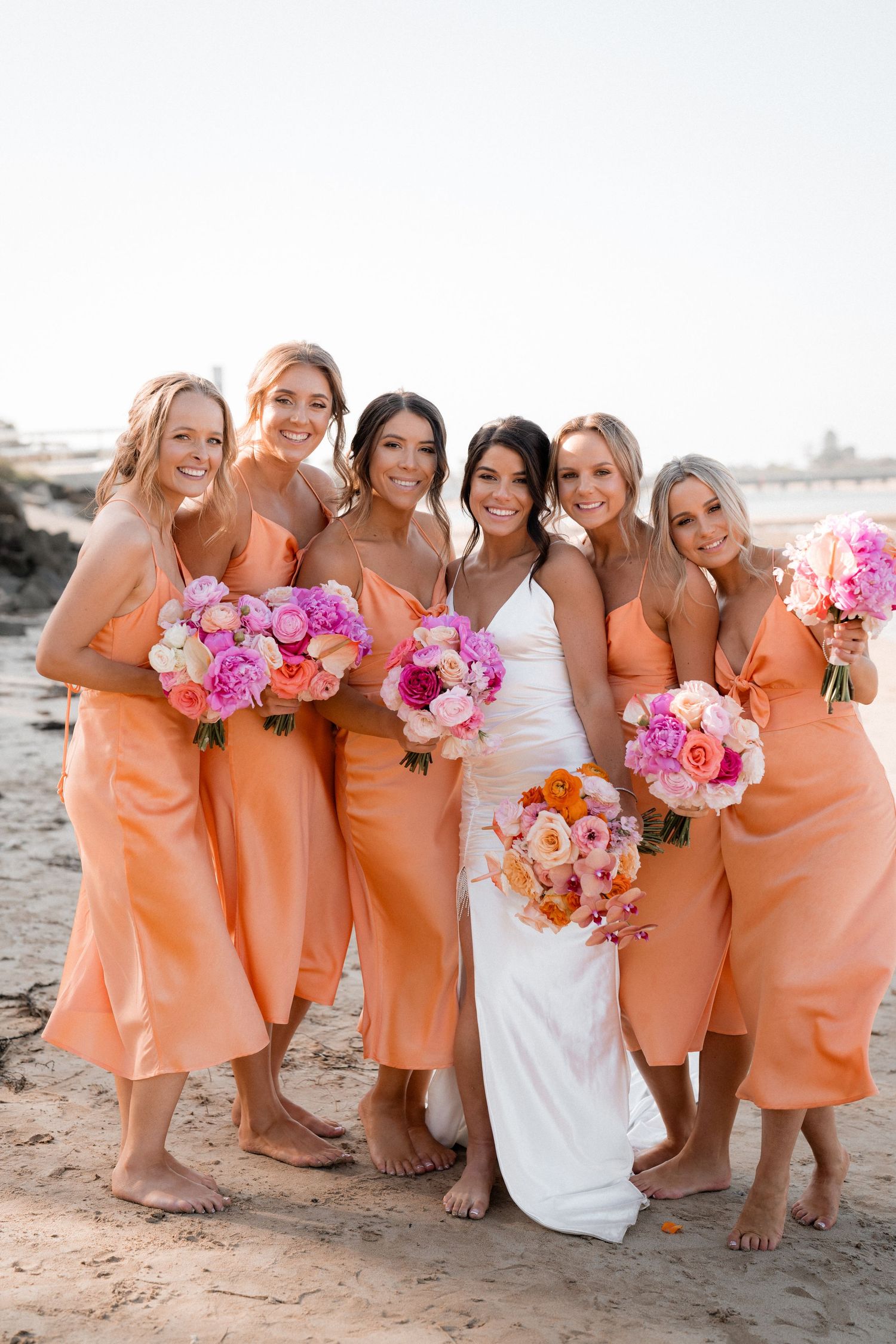 Bridal party in coral orange dresses posing together on beach with pink bouquets.