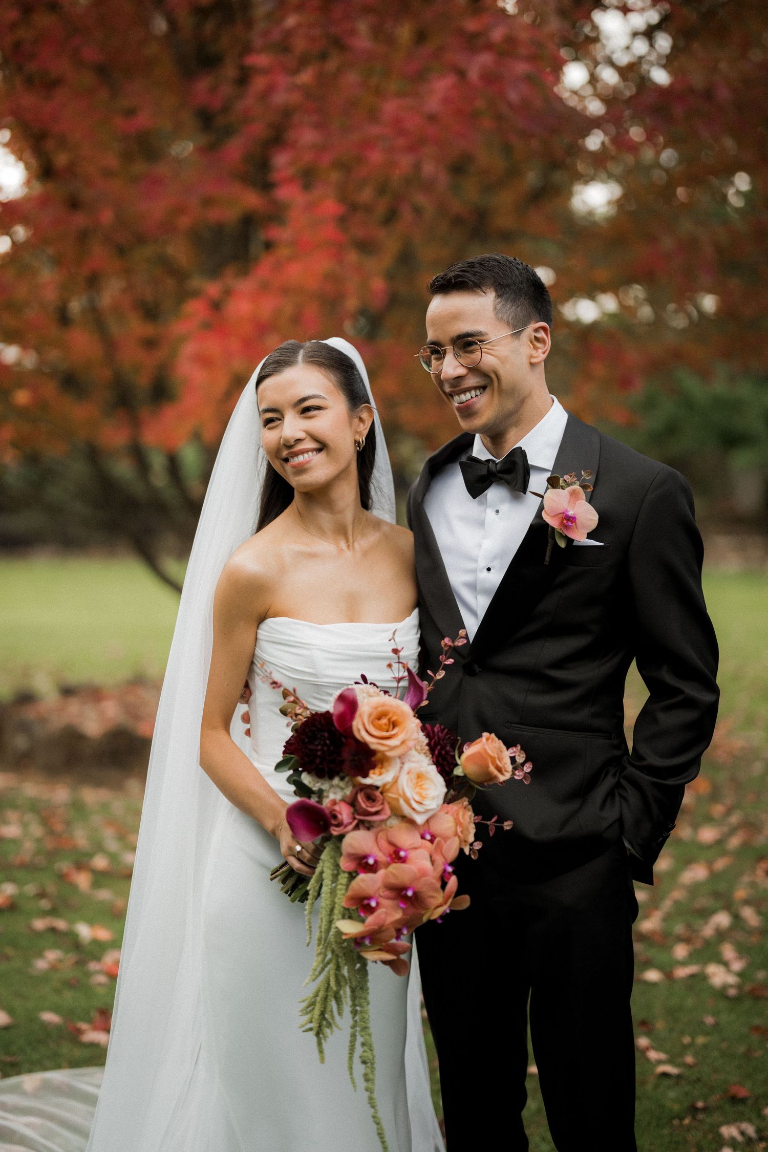 Smiling bride and groom pose outdoors in fall foliage, bride in strapless gown with colorful bouquet, groom in black tuxedo.