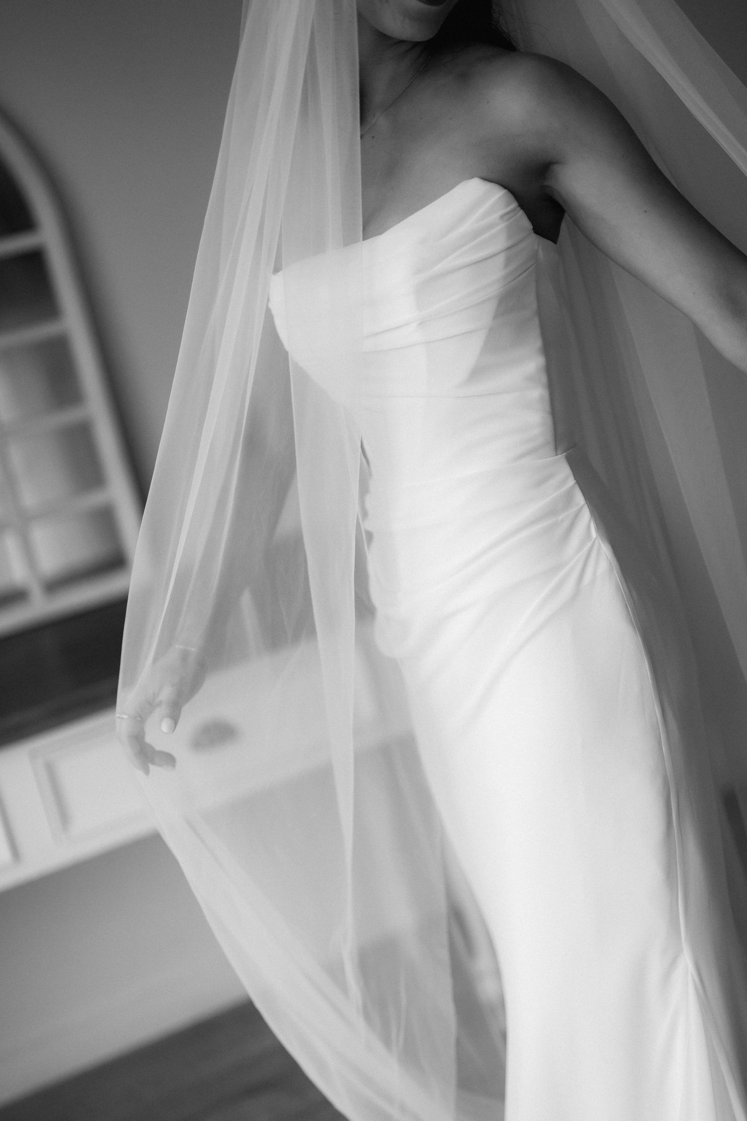 Elegant bride in strapless white satin wedding gown with flowing veil captured in dreamy black and white portrait.