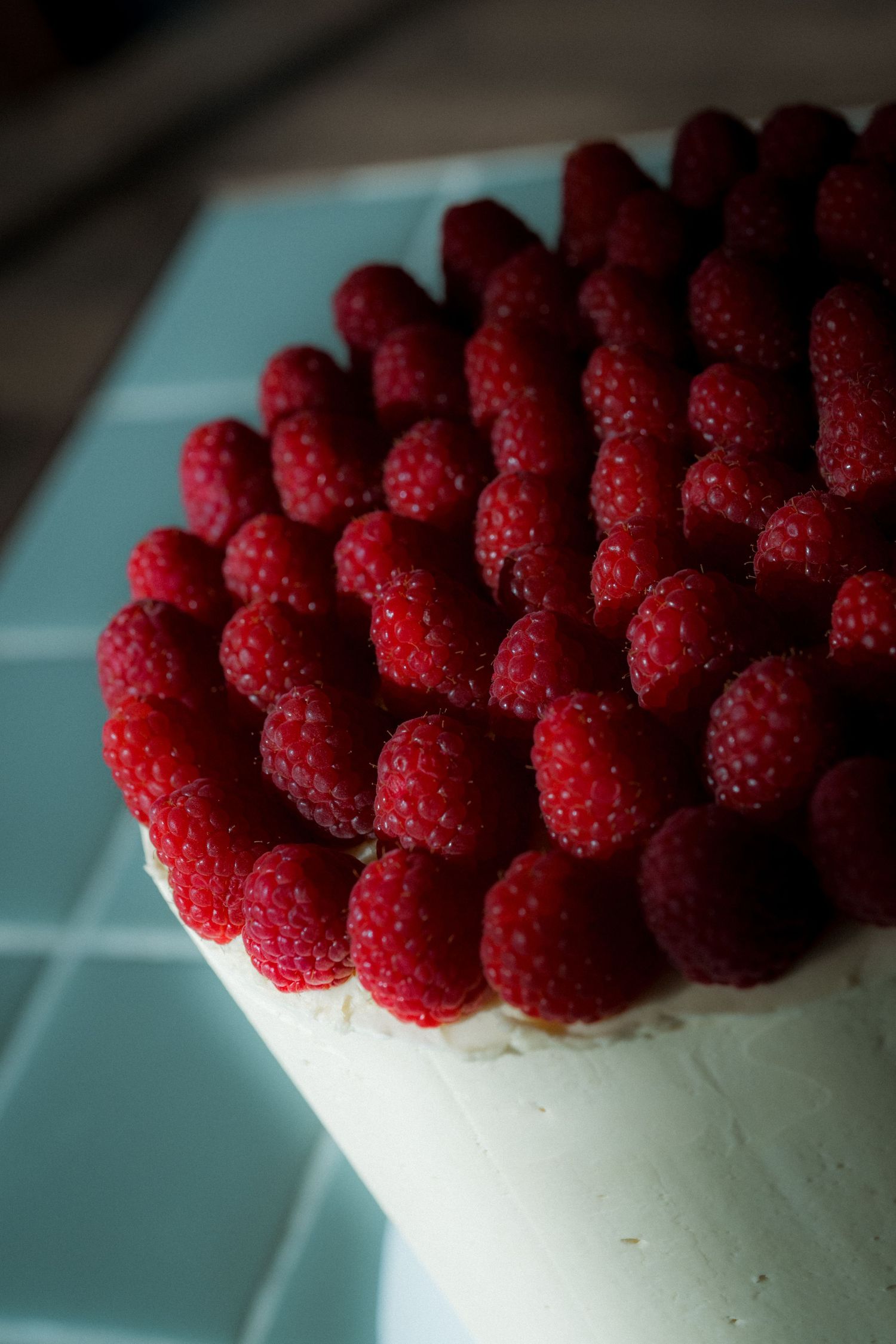 A elegant white cake topped with fresh red raspberries arranged in a dense pattern on a light blue tiled surface.