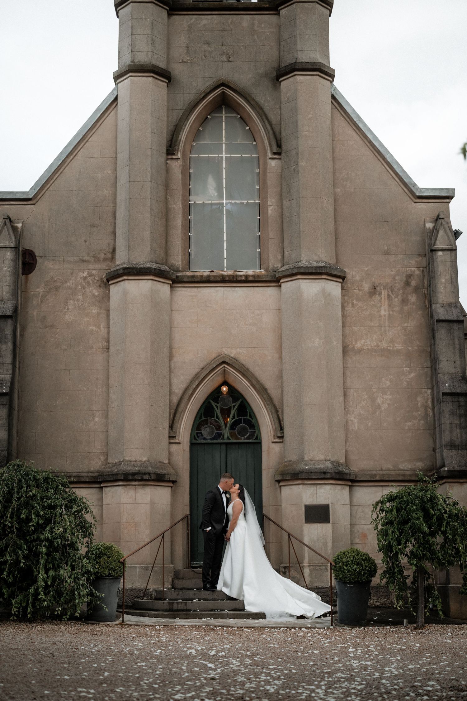 Newlywed couple standing at entrance of Gothic church with long flowing wedding dress trailing down stone steps.