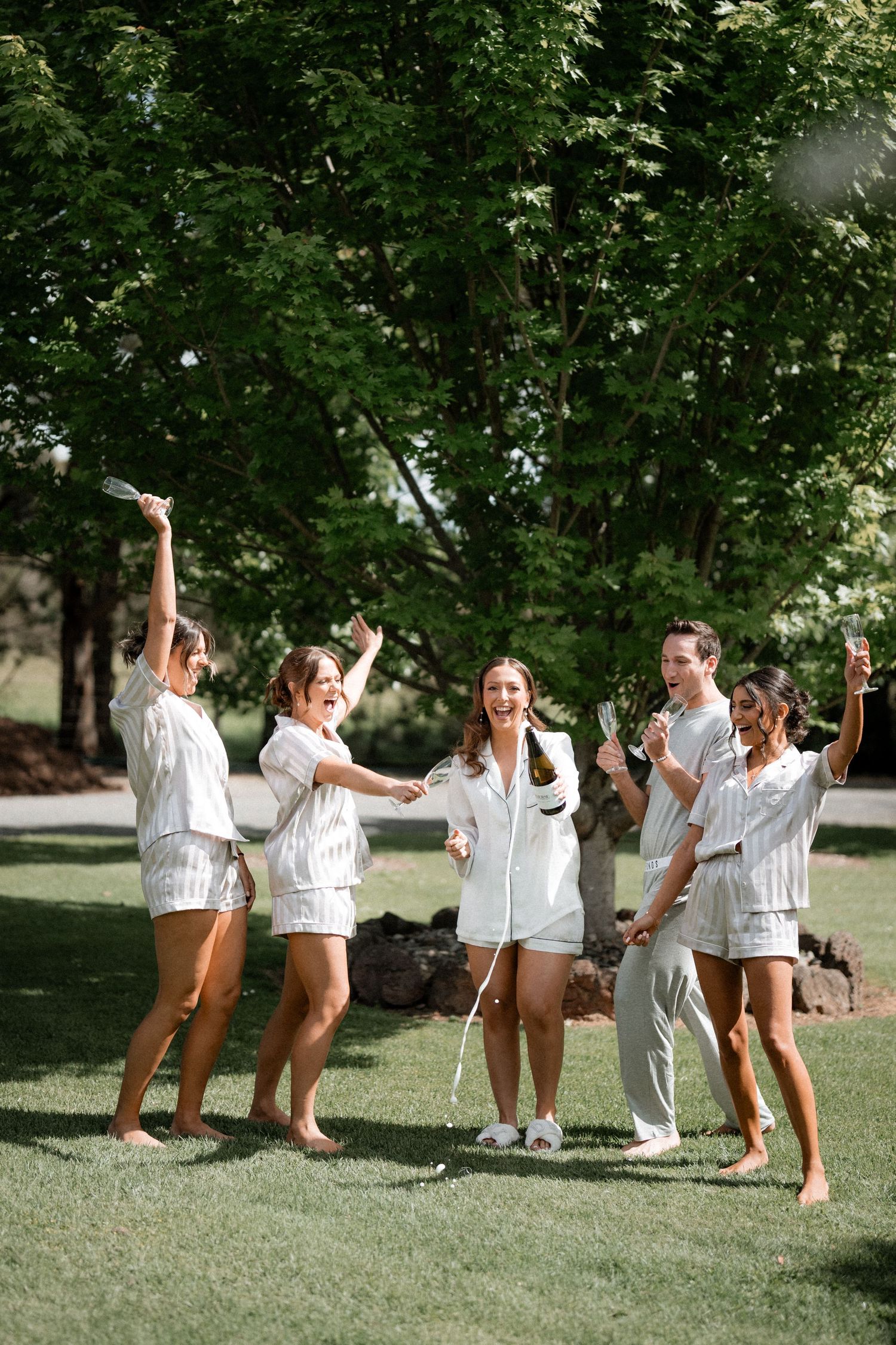 Group of friends in matching white tops and brown pants dancing joyfully together on a grassy lawn.