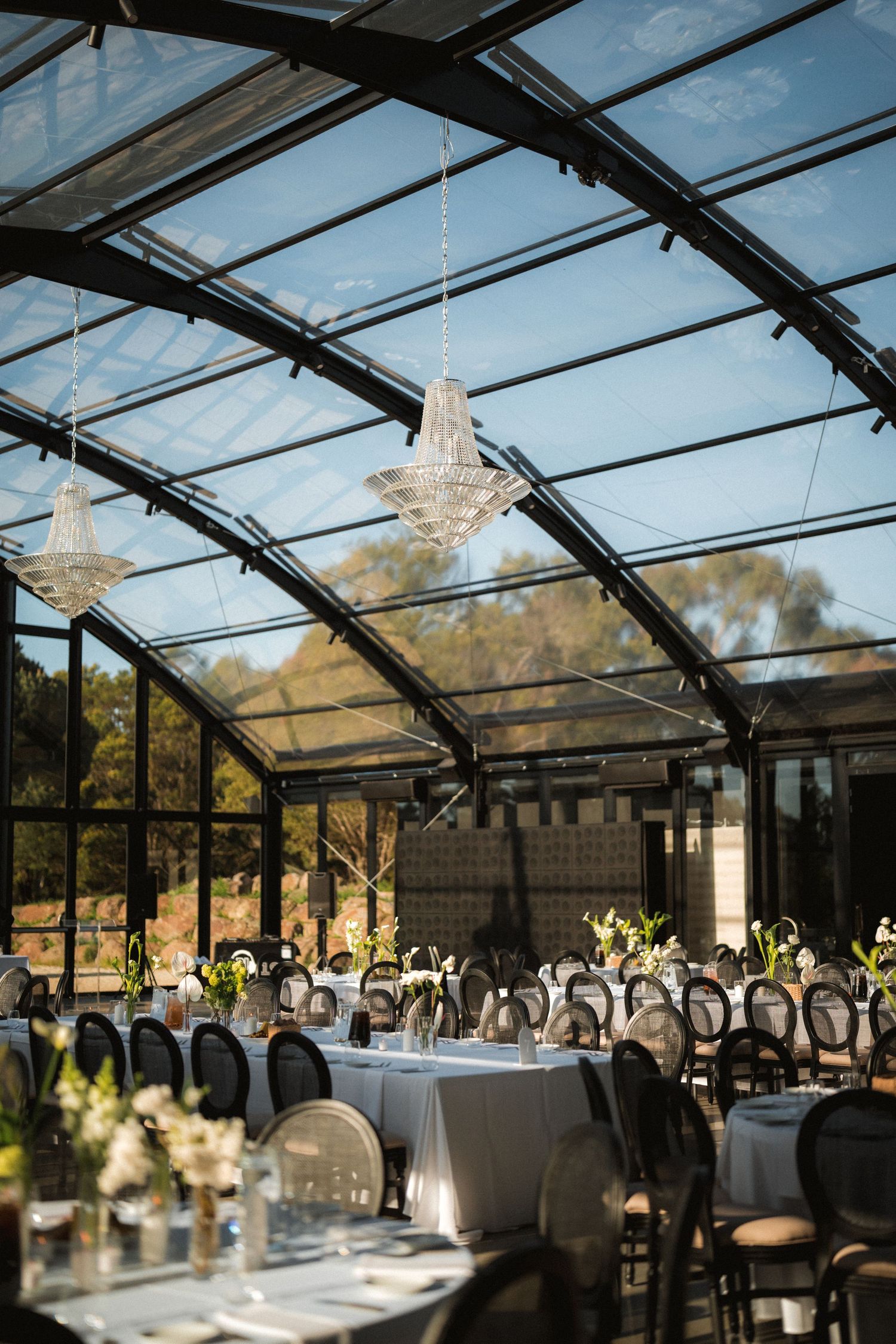 Elegant glass-roofed wedding reception venue with crystal chandeliers, round tables with white linens, floral centerpieces and garden views.