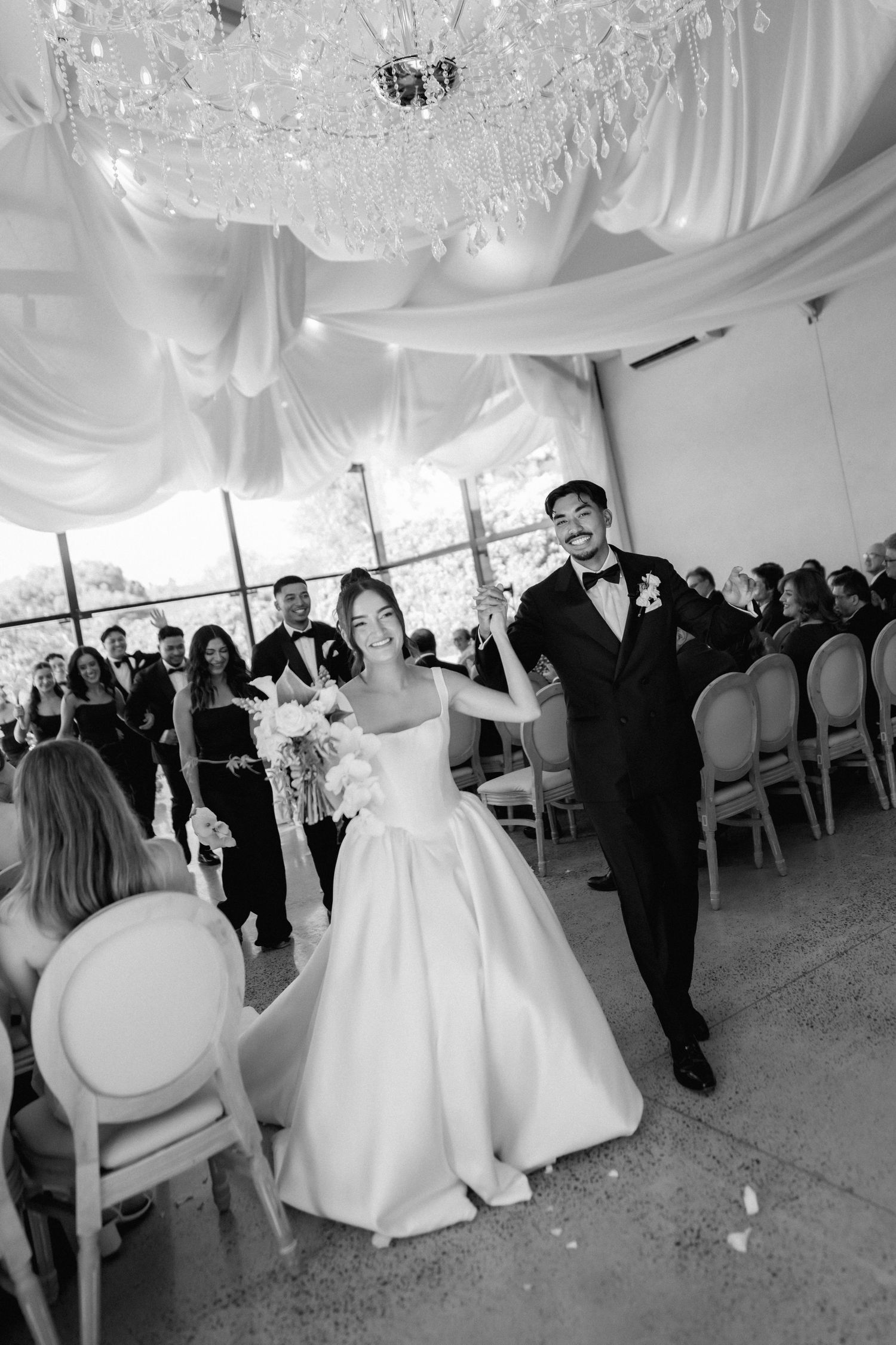 Bride in a satin gown holding a bouquet walks hand in hand with the groom down the aisle as guests cheer under a crystal chandelier.