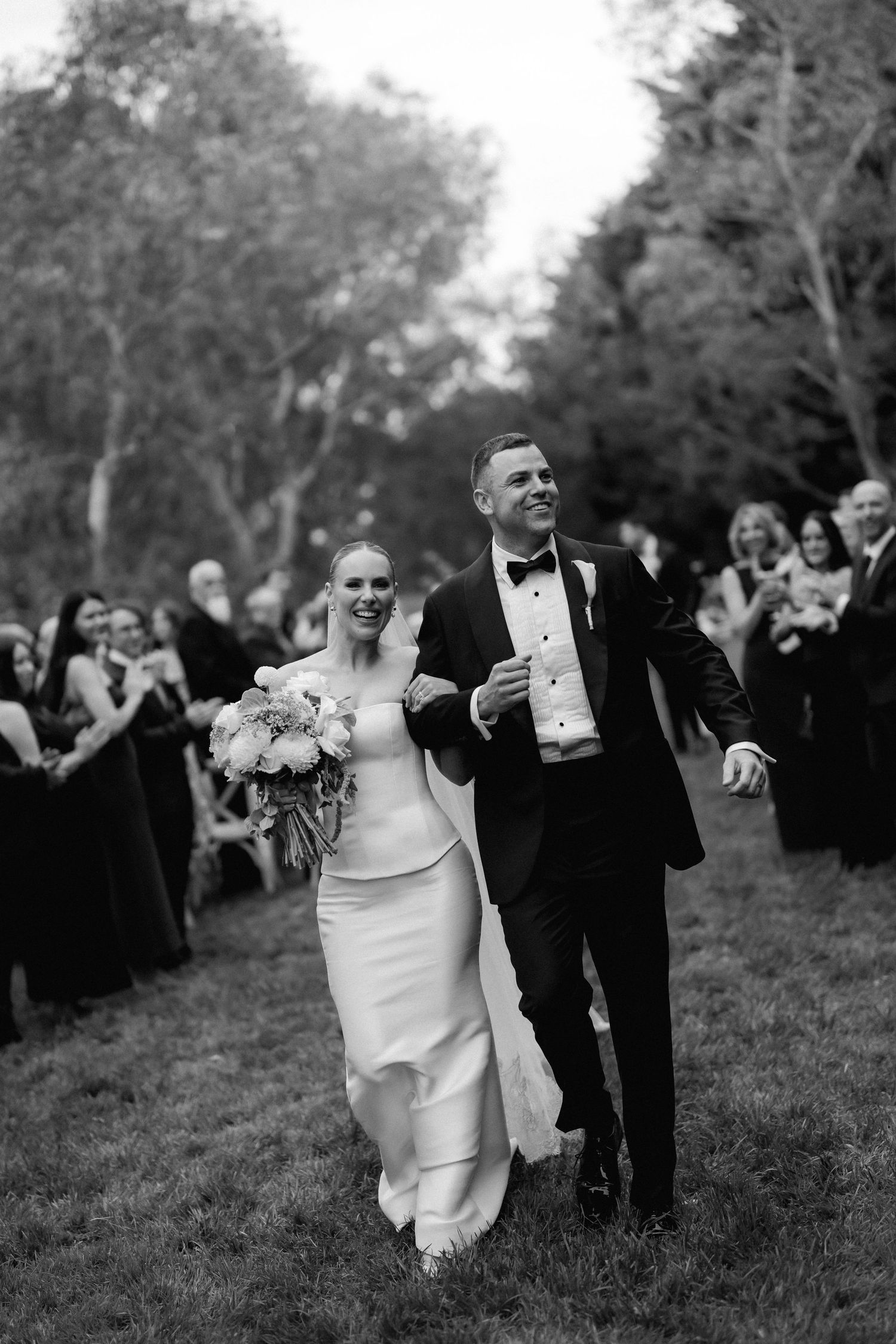 Wedding celebration on grassy lawn with guests in black dresses watching the newlyweds.