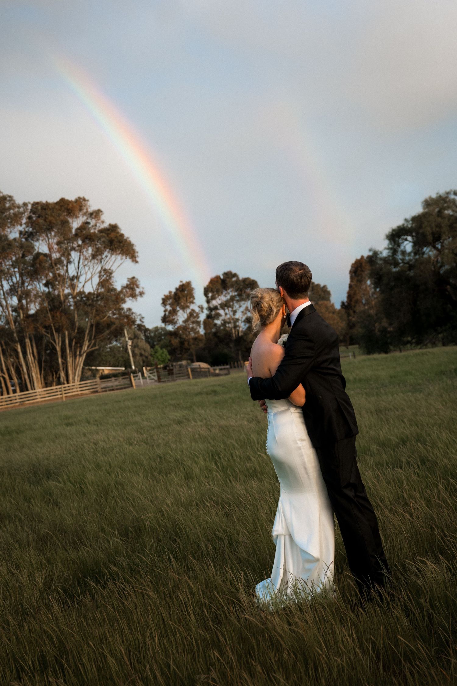 Couple embracing in a field at sunset with a rainbow appearing in the background.