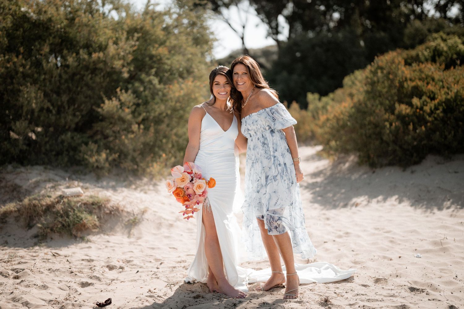 Two women in light dresses embrace on a sunny beach path while holding a bright floral bouquet.