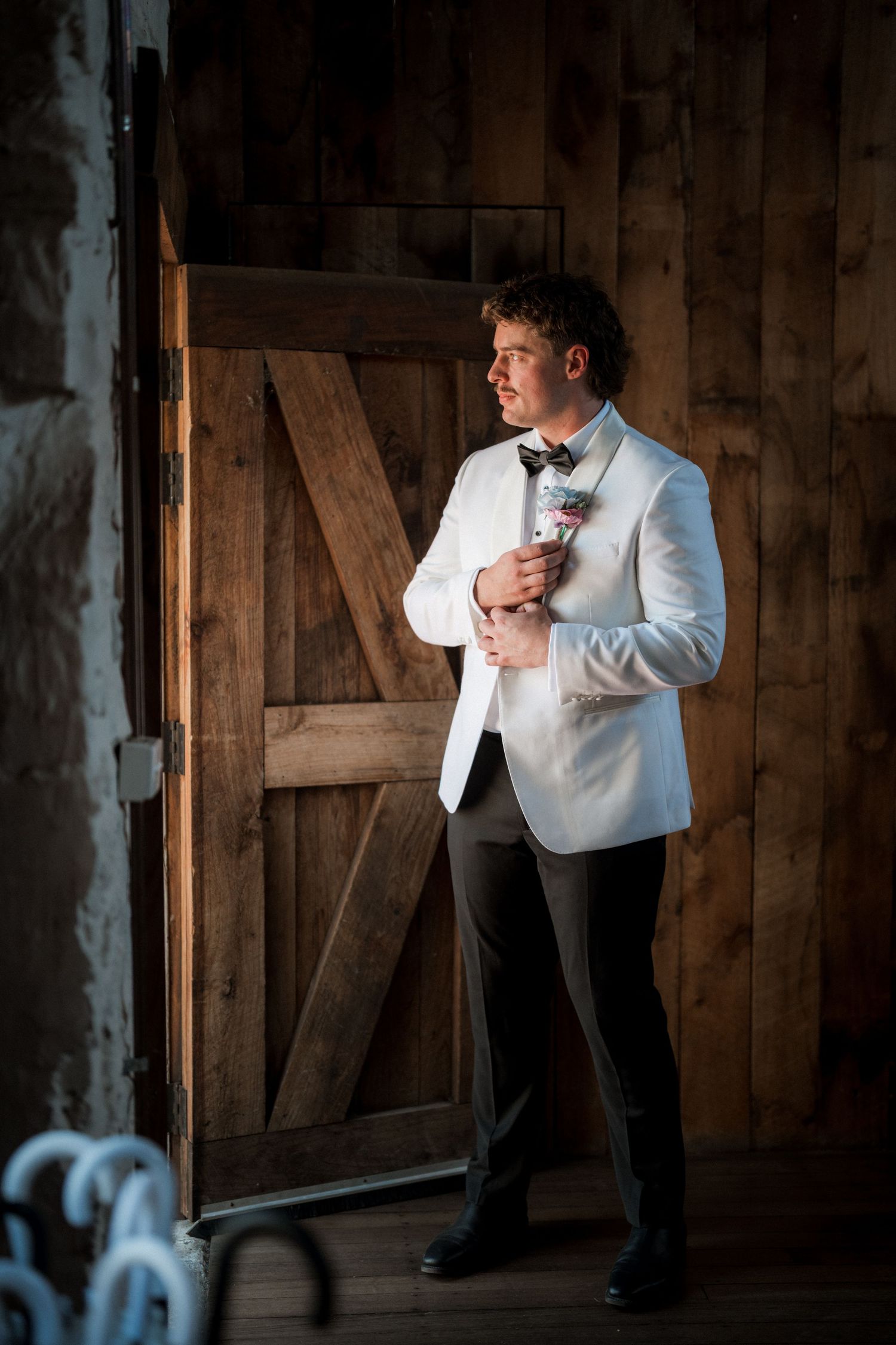 Dapper groom in white tuxedo jacket adjusting boutonniere by rustic wooden barn door.
