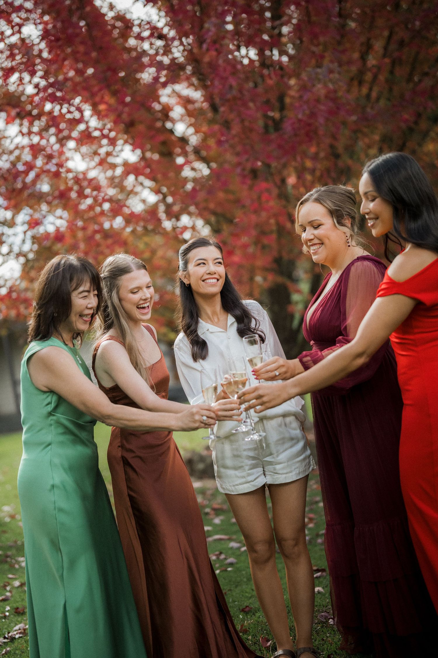 Five bridesmaids in colorful gowns toasting champagne outdoors beneath vibrant red autumn trees.