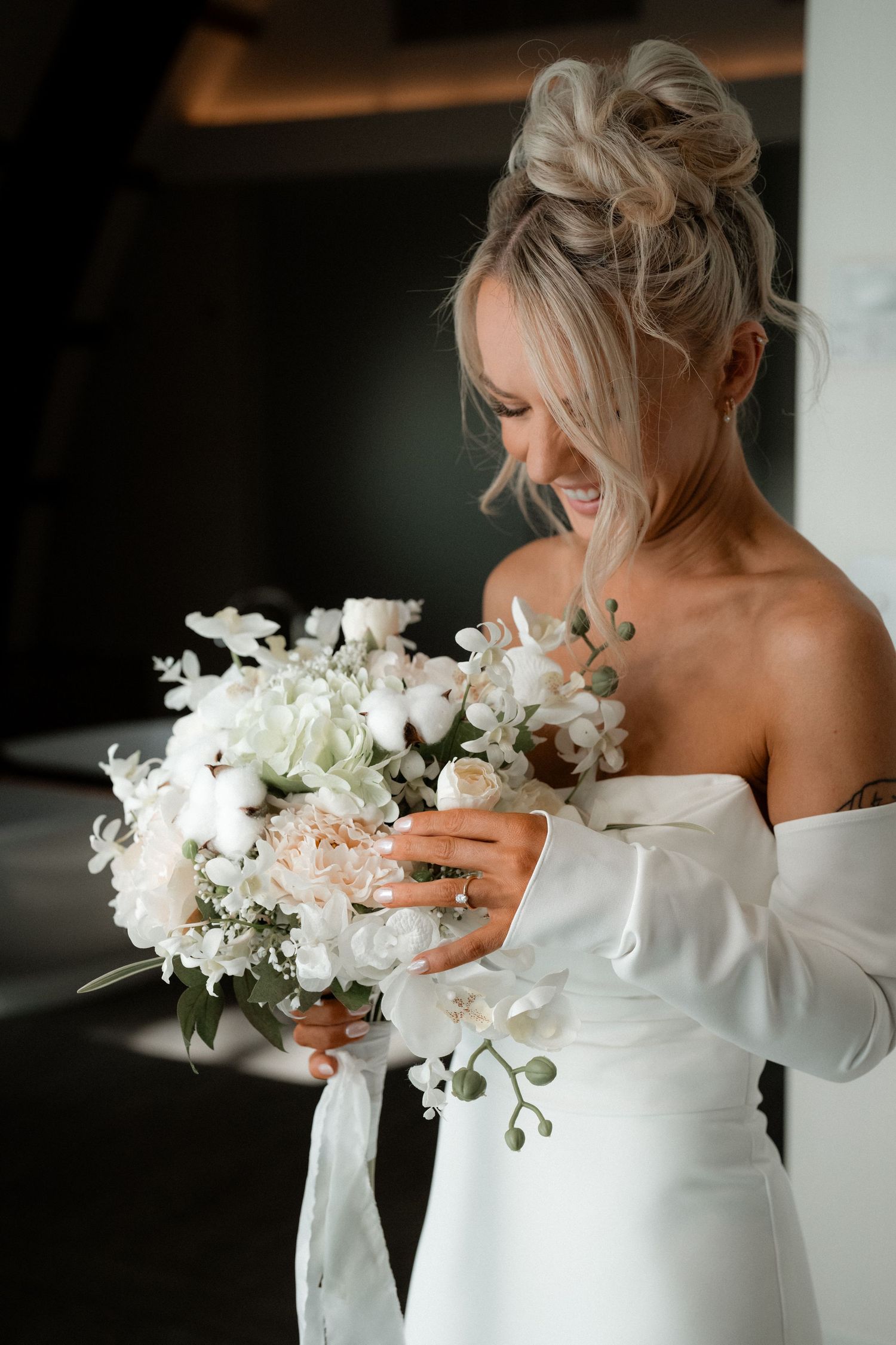 A joyful bride in a white off-shoulder dress holds a stunning bouquet of white and blush flowers with greenery.