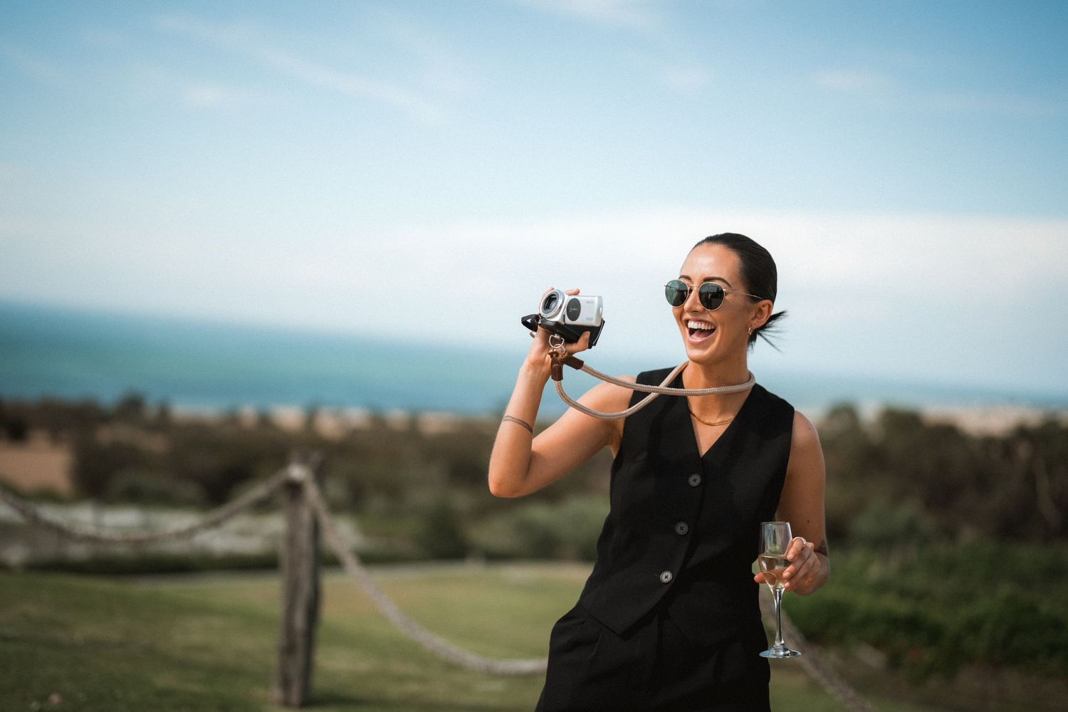 Wedding photographer in black dress holds camera and wine glass against coastal backdrop.