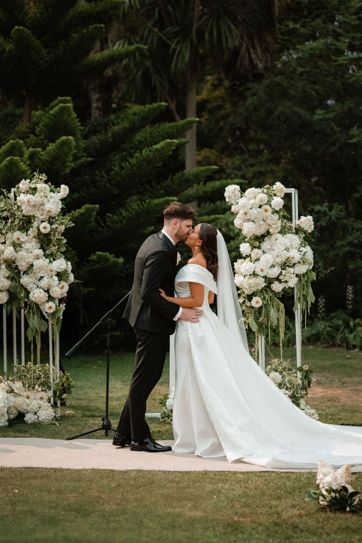 A romantic wedding ceremony under a lush garden arch adorned with elegant white floral arrangements and flowing fabric.