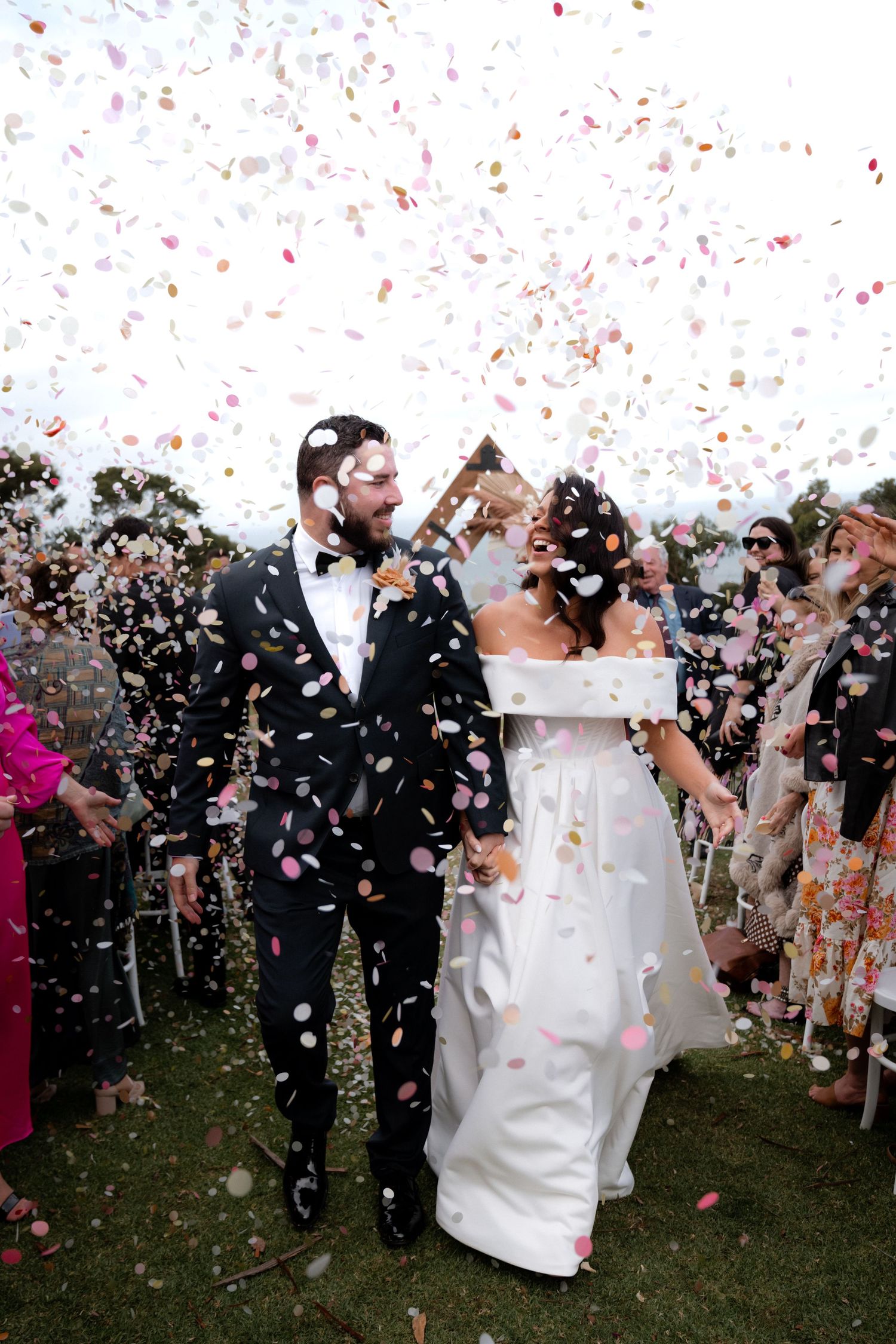 Newlyweds celebrate under a shower of confetti during an outdoor wedding ceremony with guests looking on.