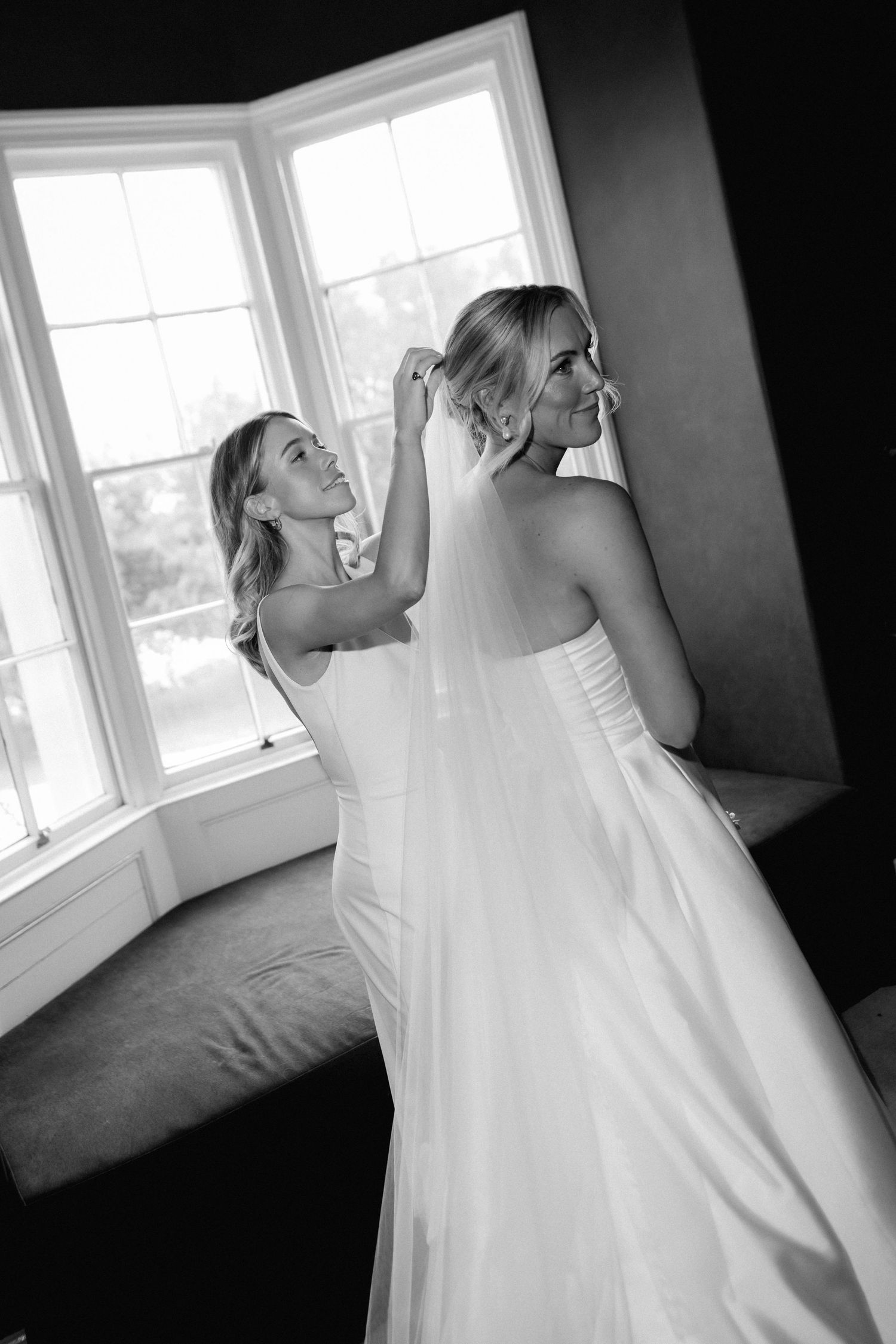 A black and white photo showing final wedding dress preparations by a bright window.